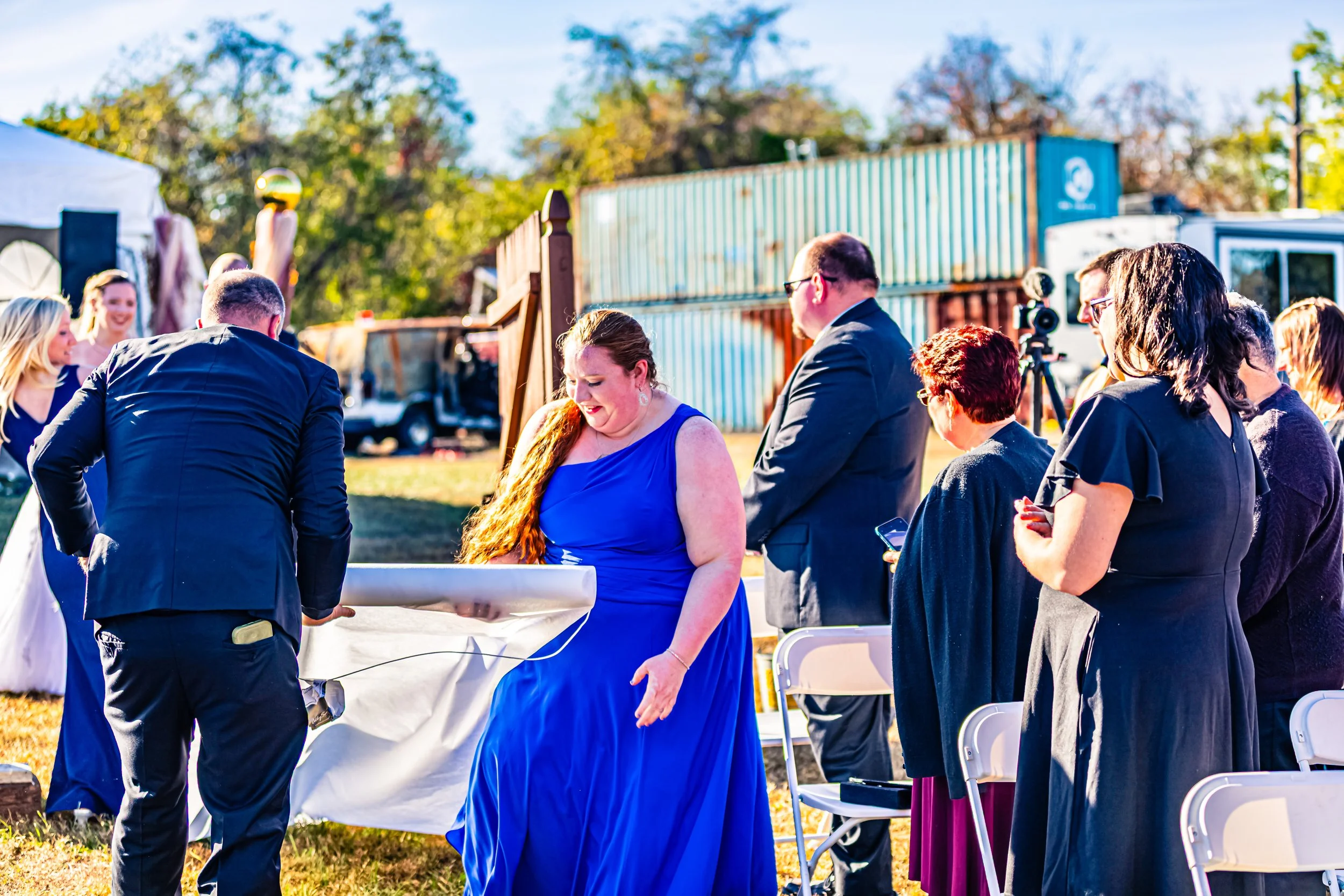 People gathered outdoors, some standing and some sitting, during a joyful celebration, with a woman in a bright blue dress at the center.