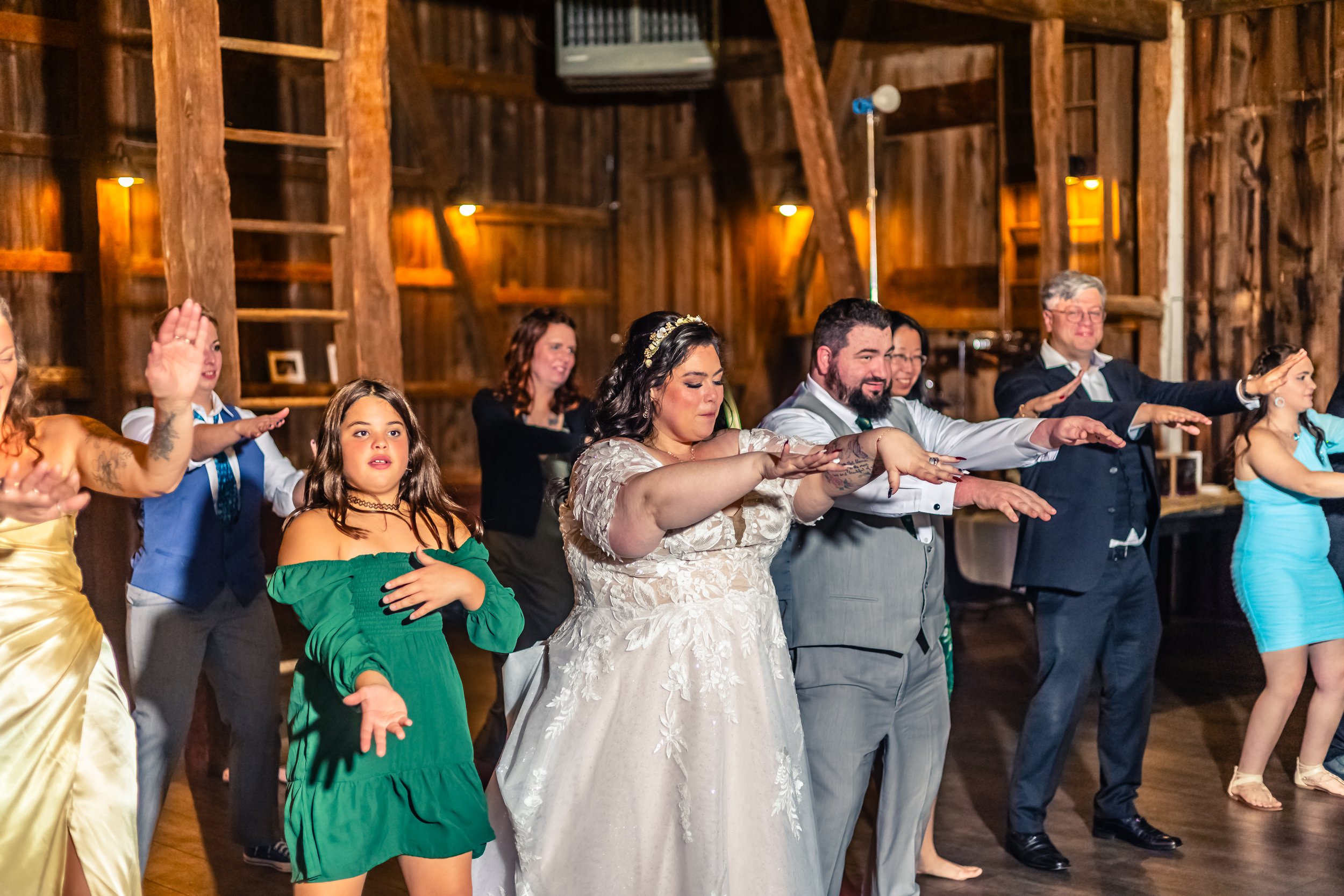 A group of people, including a bride in a wedding dress, performing a dance together in a rustic wooden venue, with arms extended forward and diverse expressions.