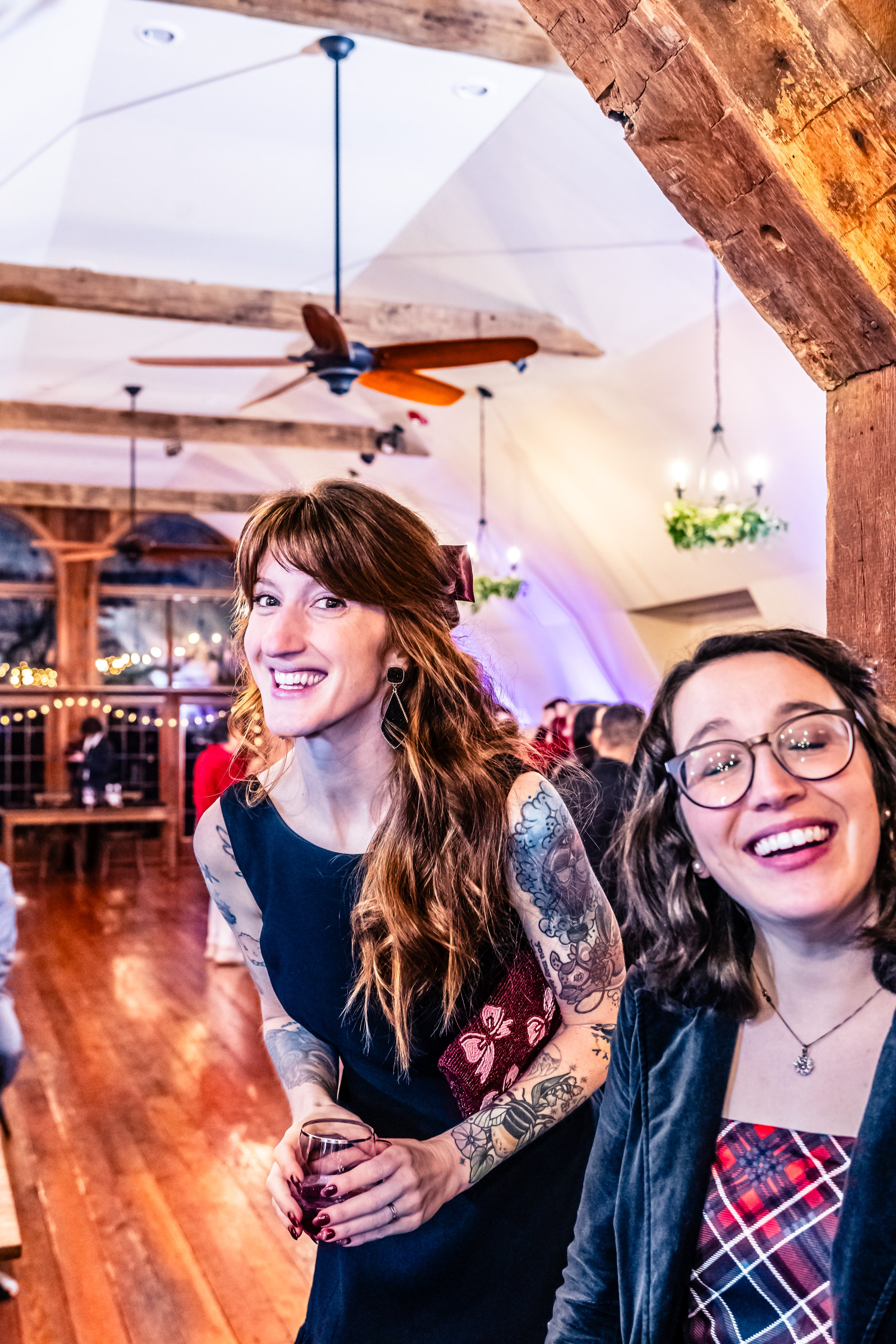 Two women smiling and laughing at a social gathering in a warmly lit room with wooden beams and ceiling fans.