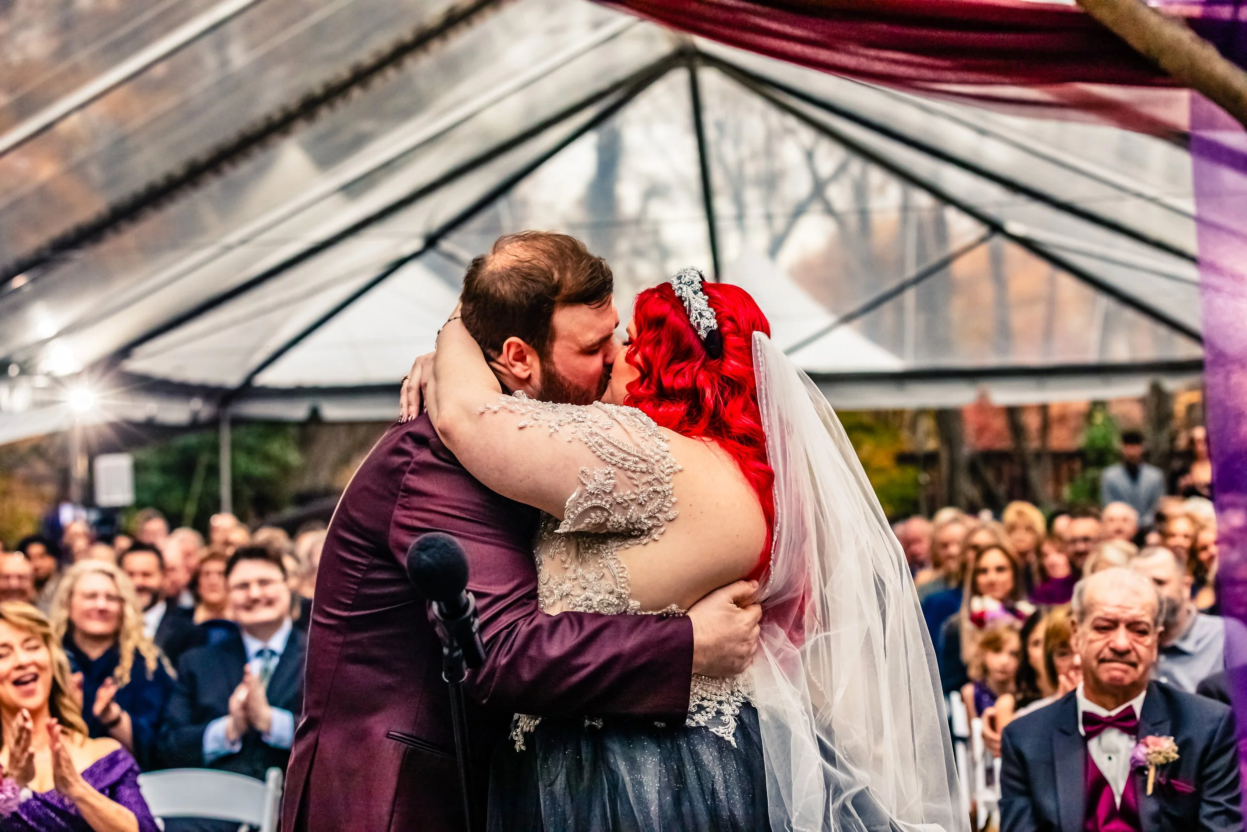 Couple kissing at wedding ceremony underneath clear tent with guests watching and clapping, autumn trees in background.