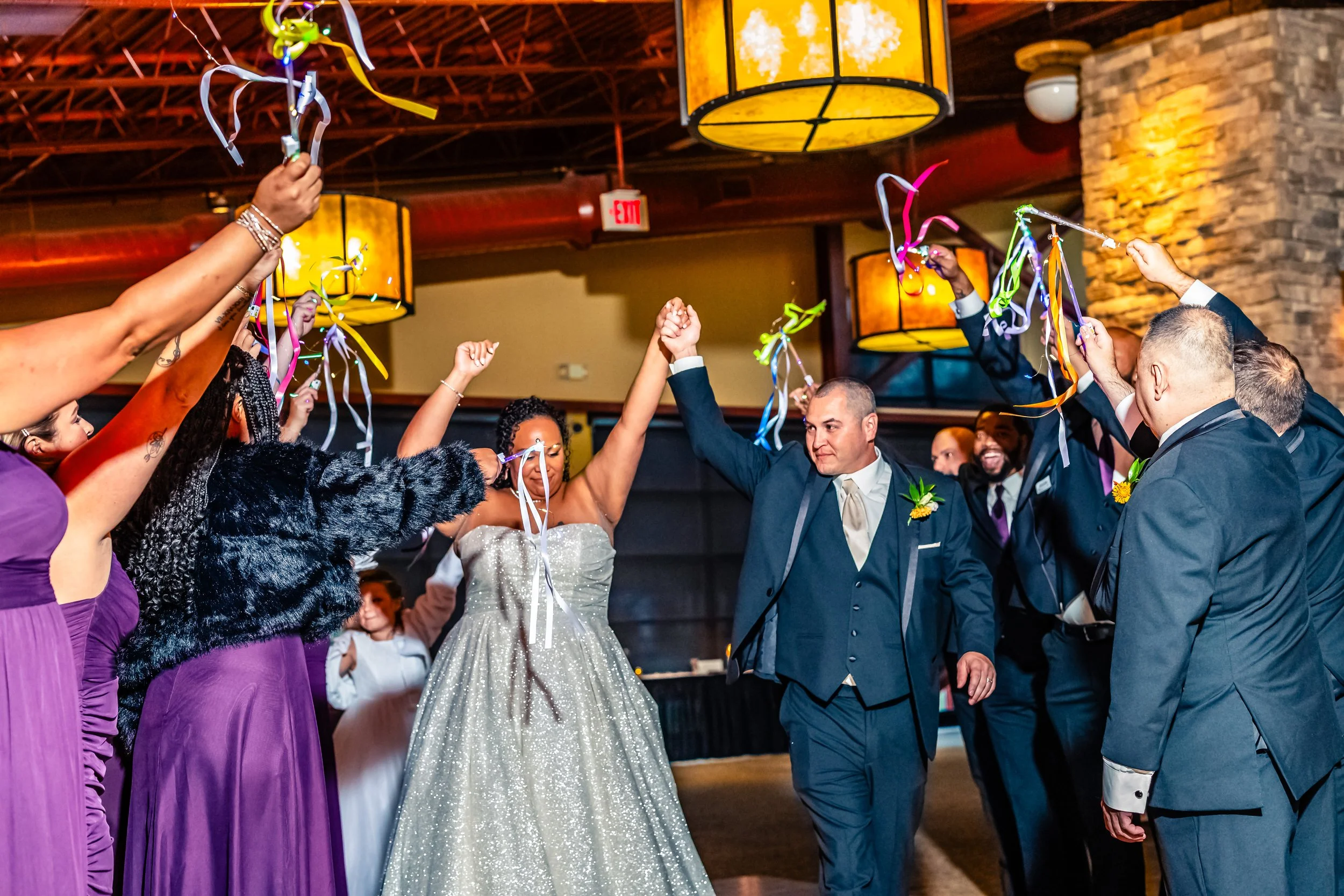 Bride and groom holding hands and raising their arms in celebration with wedding guests surrounding them, colorful streamers in the air at wedding reception.