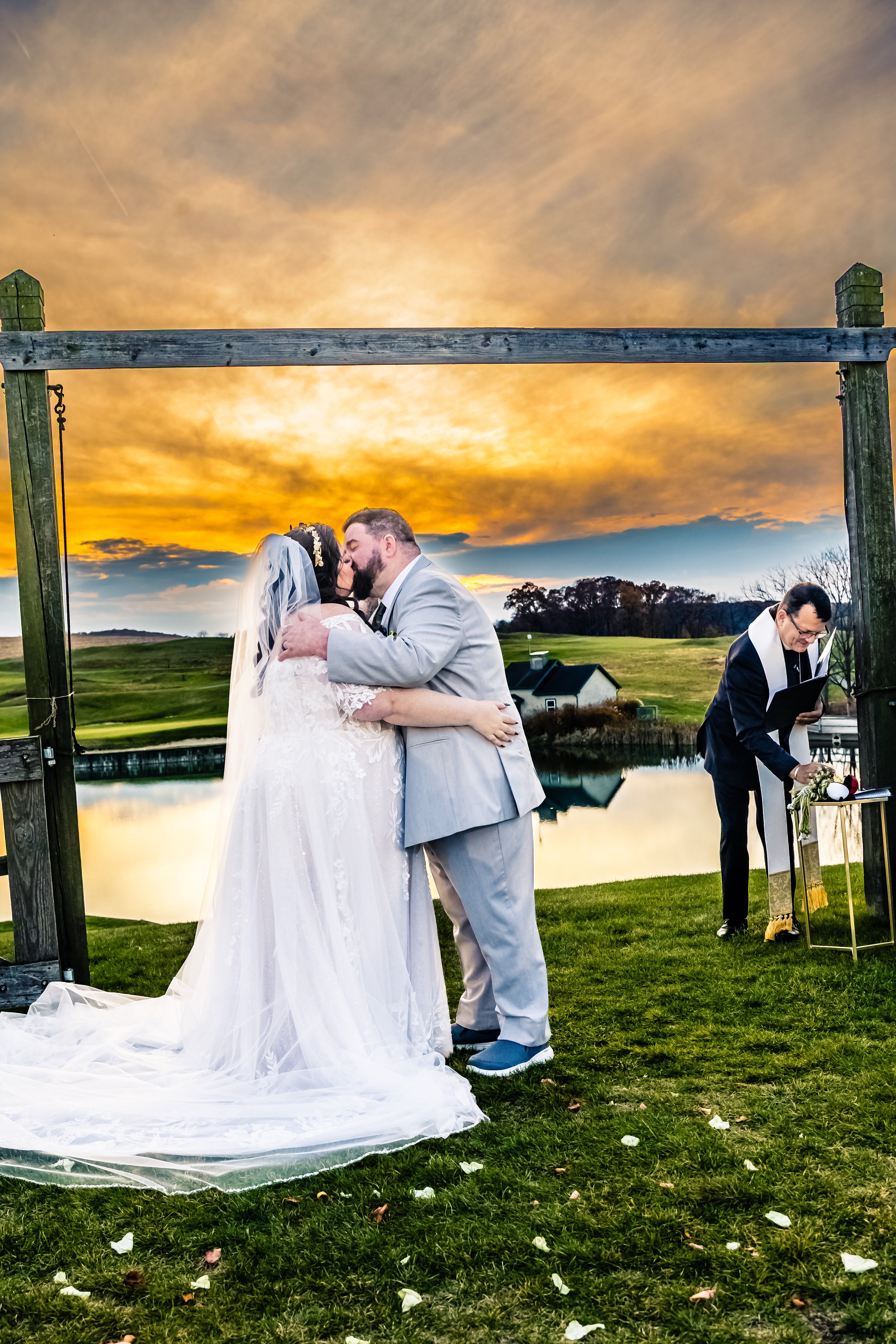 A bride and groom kiss during their outdoor wedding ceremony at sunset by a lake, with a pastor officiating and a scenic rural landscape in the background.