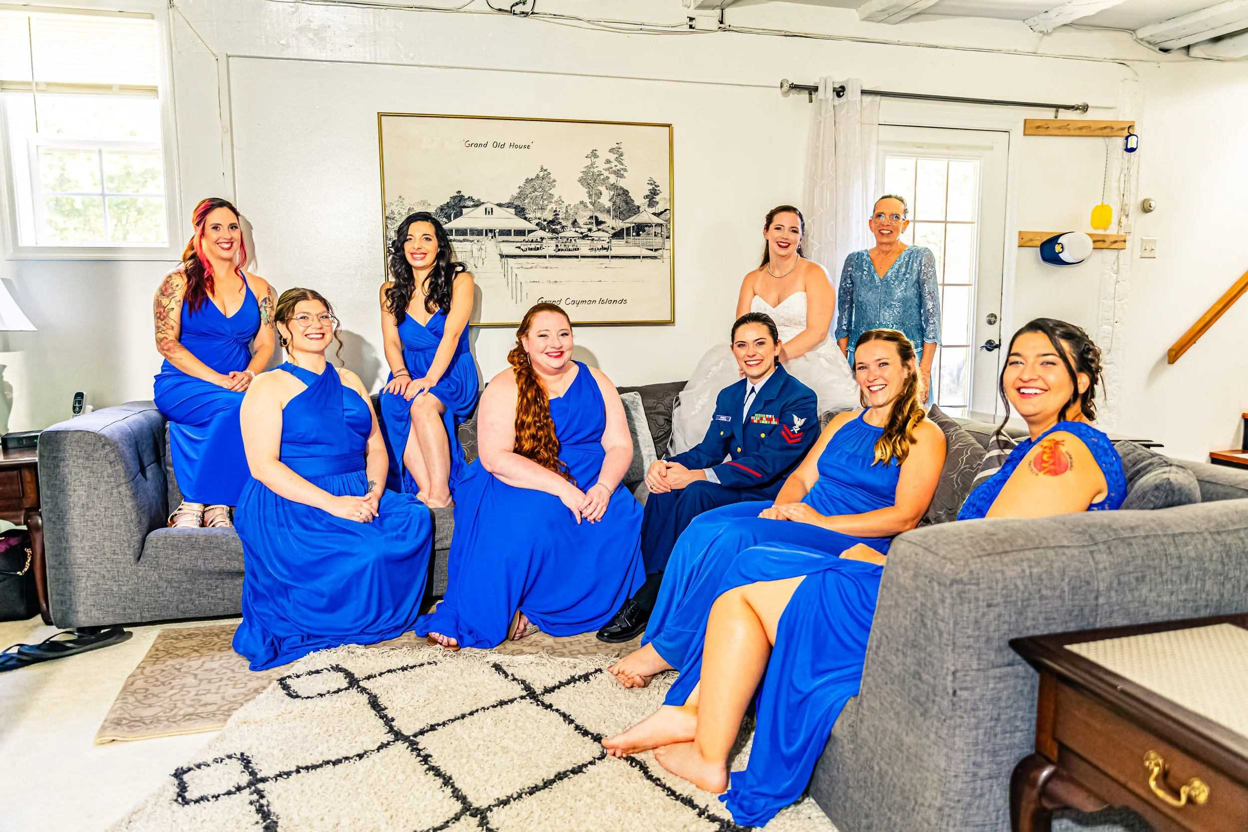 A group of nine women, some in bridesmaids dresses and one in a military uniform, gathered in a living room. They are smiling and sitting on and around a gray couch, with a bride in a white wedding gown standing behind them. The room has white walls,