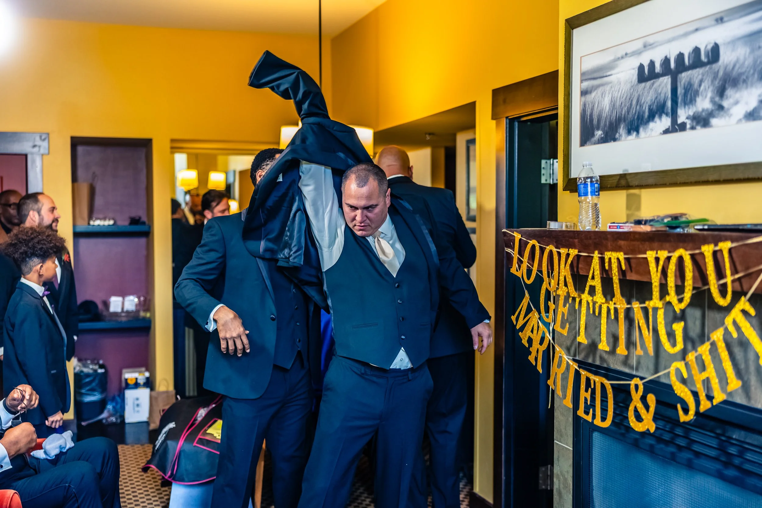 A group of men and boys dressed formally gather inside a decorated room with yellow walls. One man appears to be putting on a jacket, with a birthday banner that reads 'LOOK AT YOU GETTING MARRIED & SHIT' hanging near the fireplace. The scene suggest