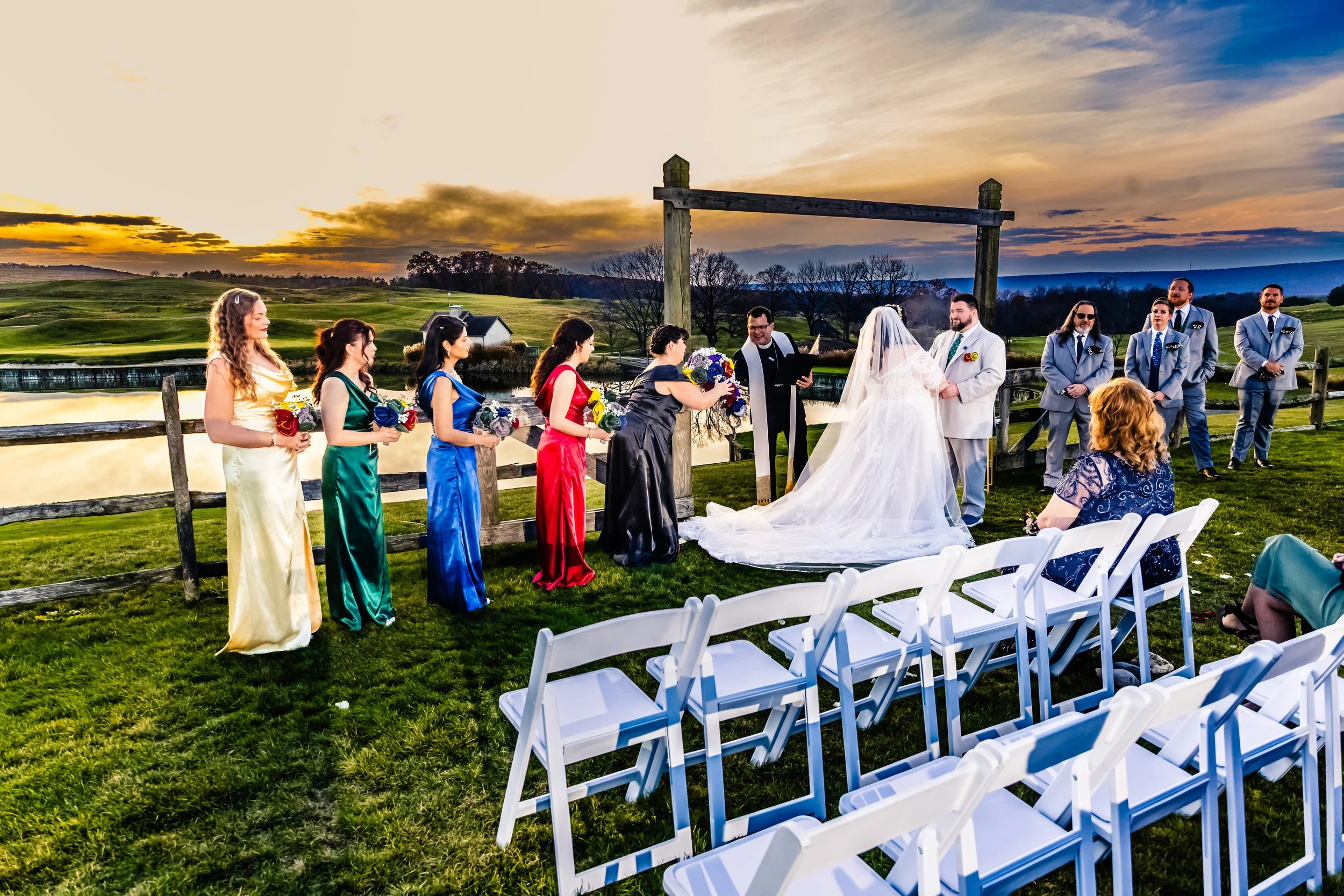 Outdoor wedding ceremony at sunset on a grassy field near a pond, with a bride and groom standing under a wooden arch, surrounded by bridesmaids and groomsmen, and a woman sitting on white chairs watching.