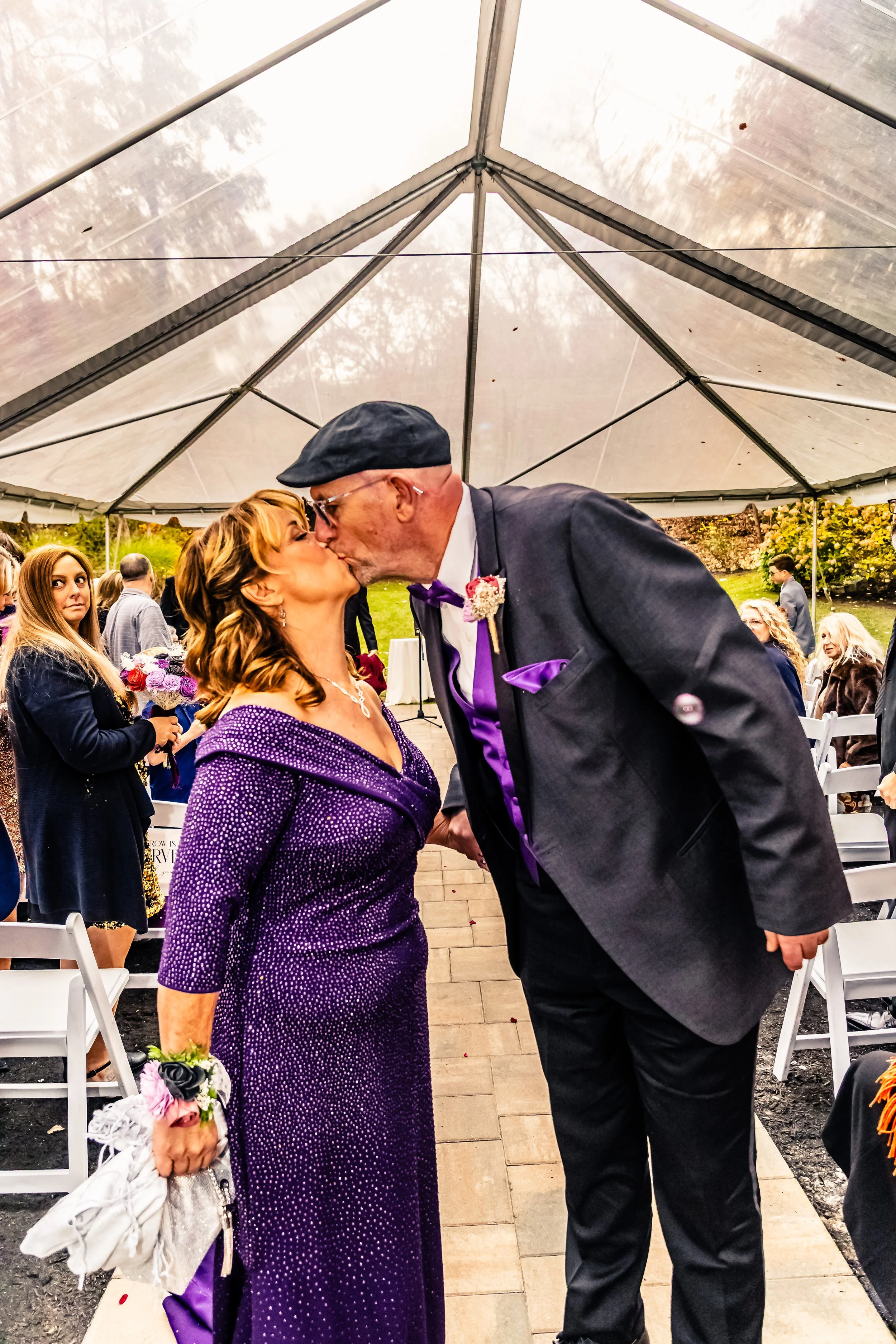 A man and woman share a kiss under a white event tent, surrounded by seated guests, at an outdoor wedding reception.