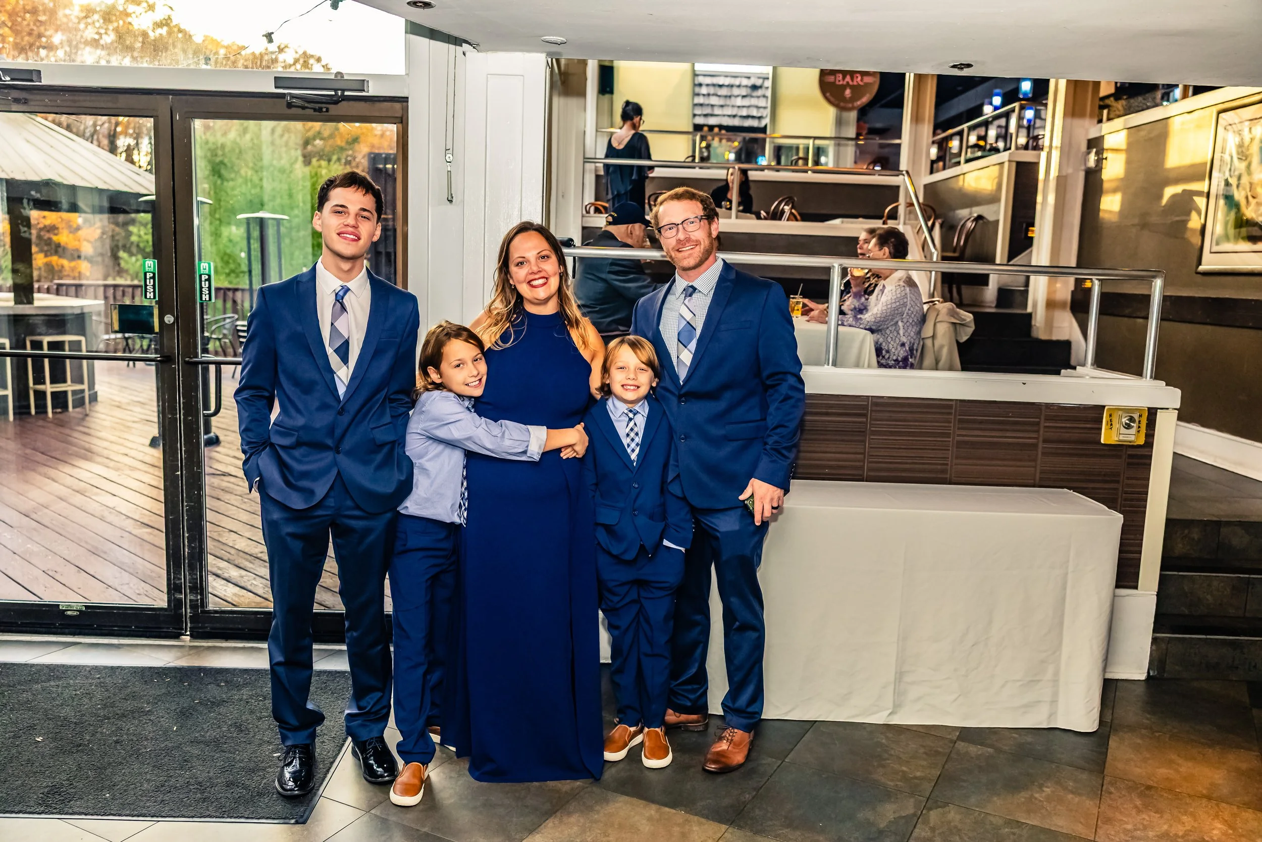 A family of five dressed in blue suits and dresses stands inside a restaurant near the entrance, smiling at the camera.