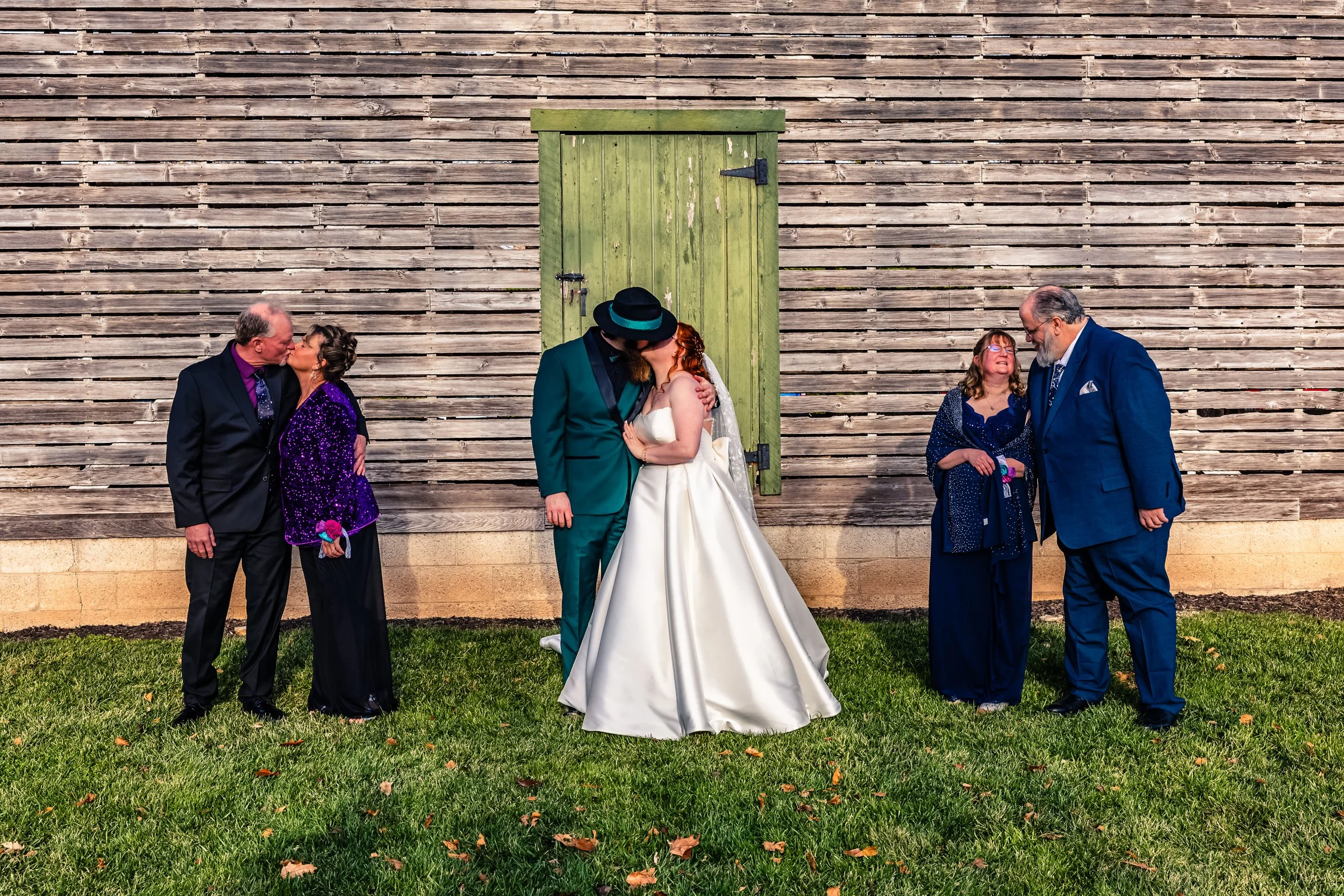 A wedding photo with two couples kissing in front of a wooden wall with a green door. The couple in the center, the bride in a white gown and the groom in a teal suit, are sharing a kiss. The other couple, on the left, is also kissing, and the couple