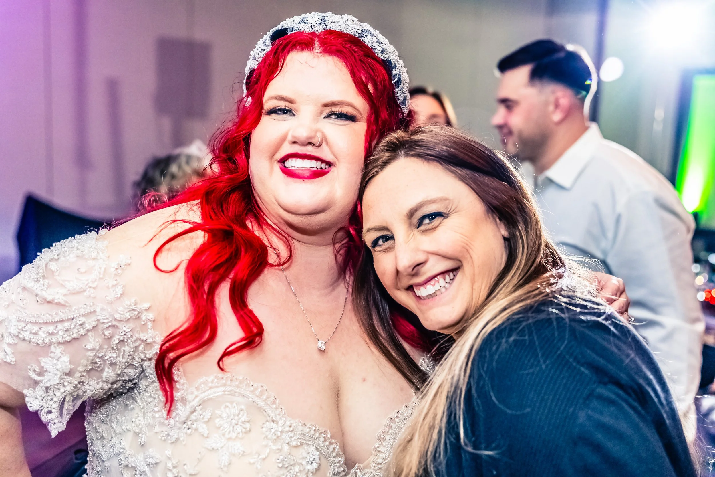 Two women smiling and hugging at a celebration, one with bright red hair and a wedding dress, the other with long brown hair and a black outfit, with blurred people in the background.