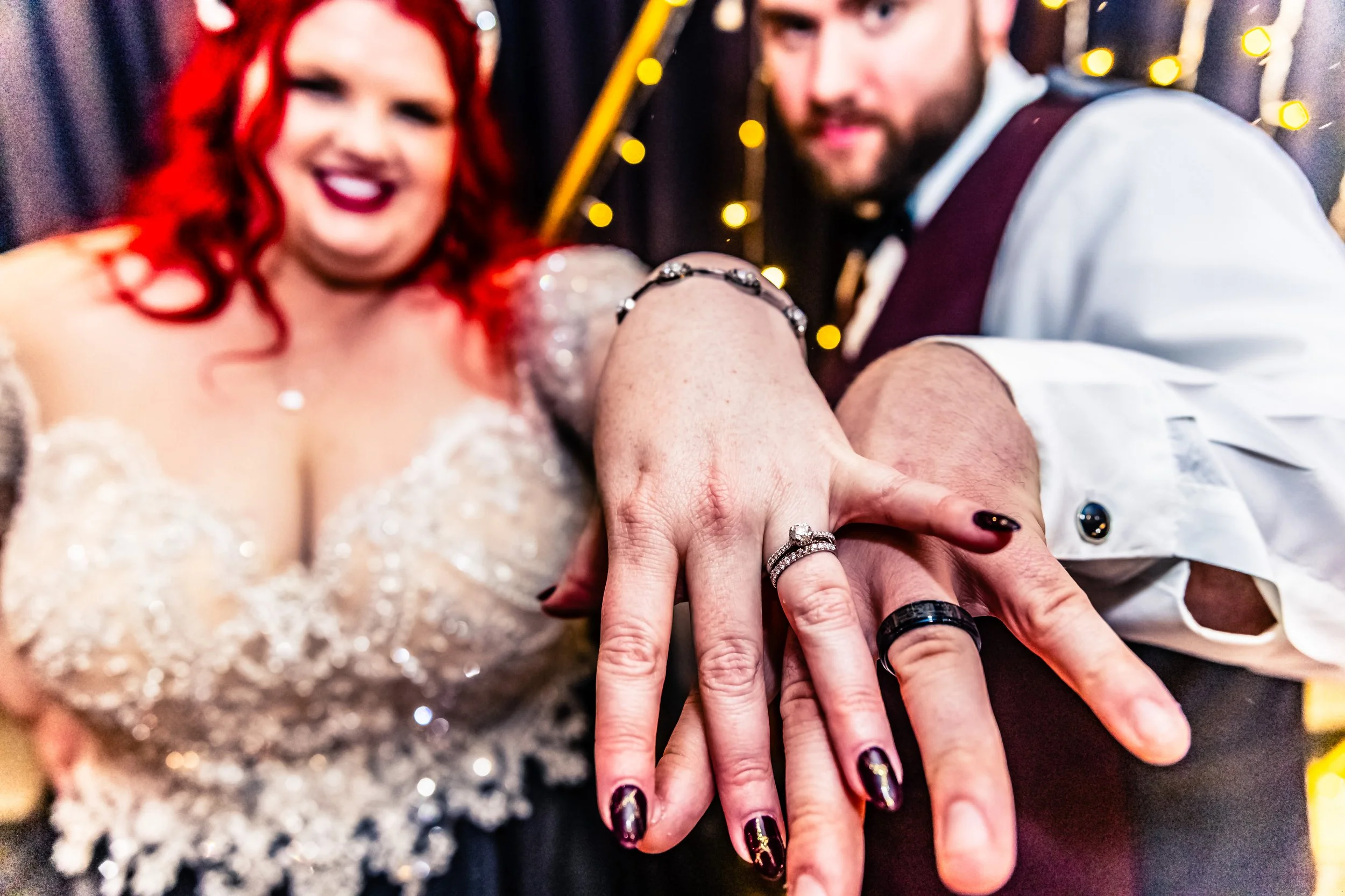 Bride and groom showing their wedding rings, smiling, with blurred party background.