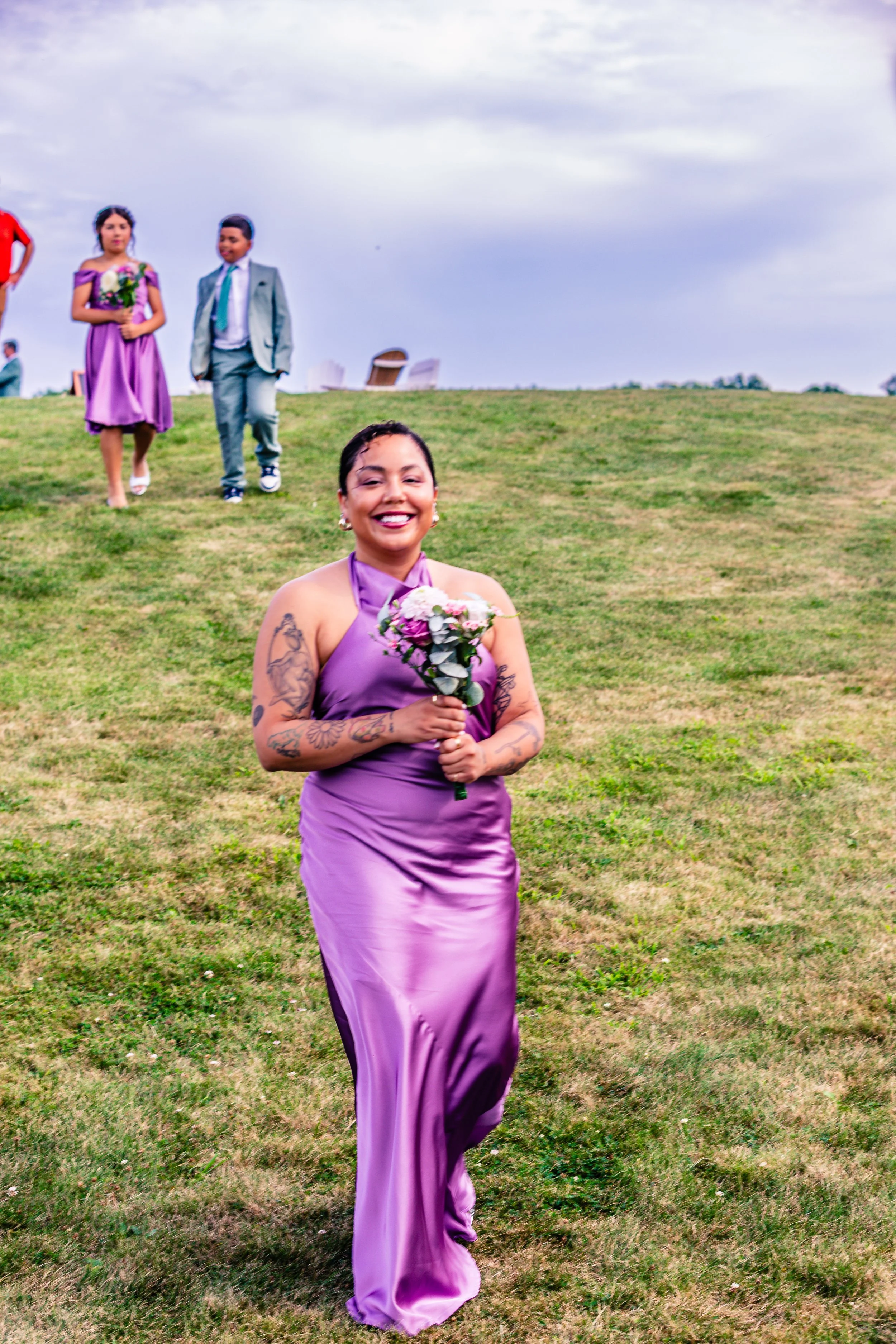 A woman in a purple satin dress holding a bouquet of flowers, smiling happily while walking on a grassy hill with other well-dressed people in the background.