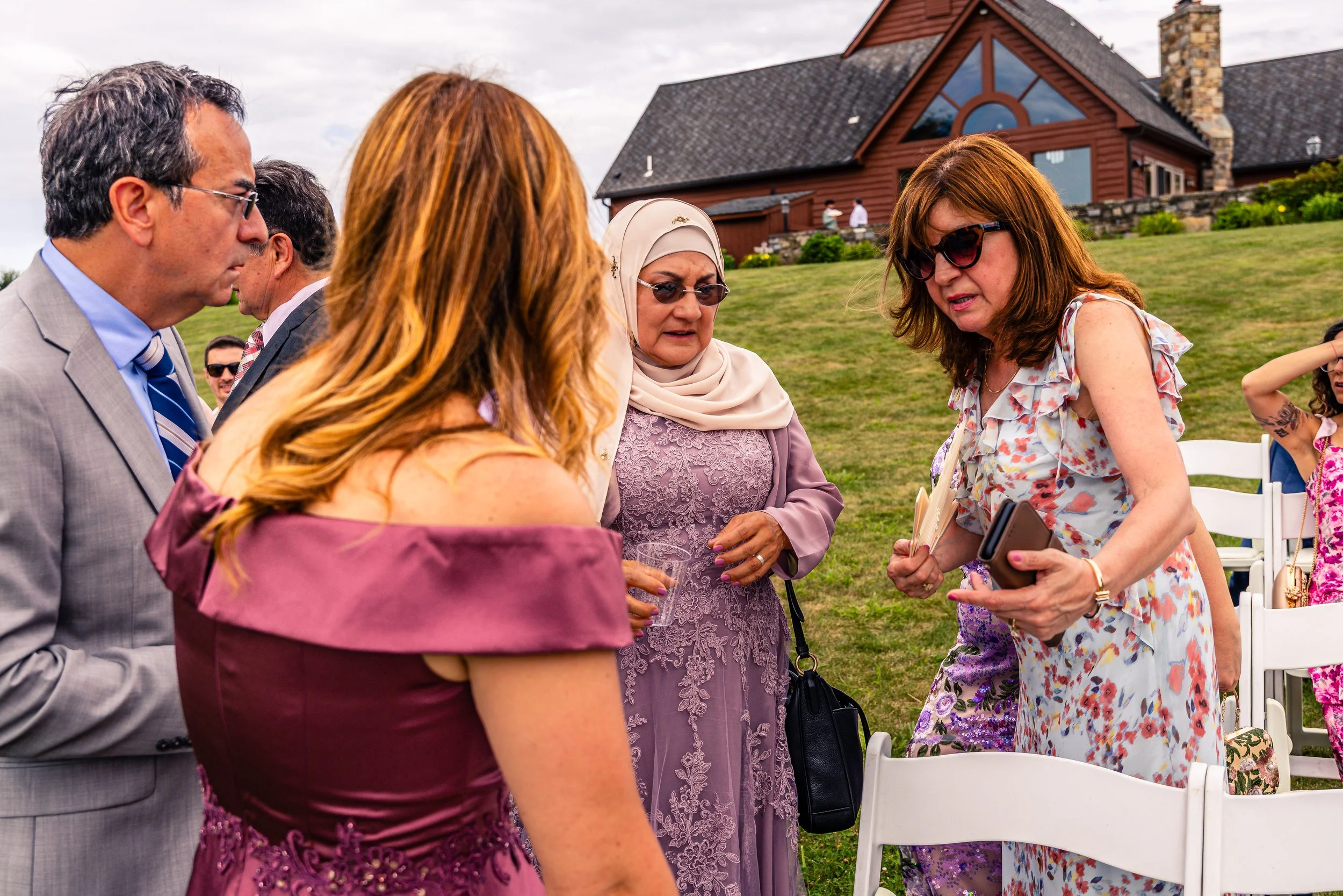 Group of people in formal attire at an outdoor event, conversing near white chairs on a grassy lawn with a large house in the background.