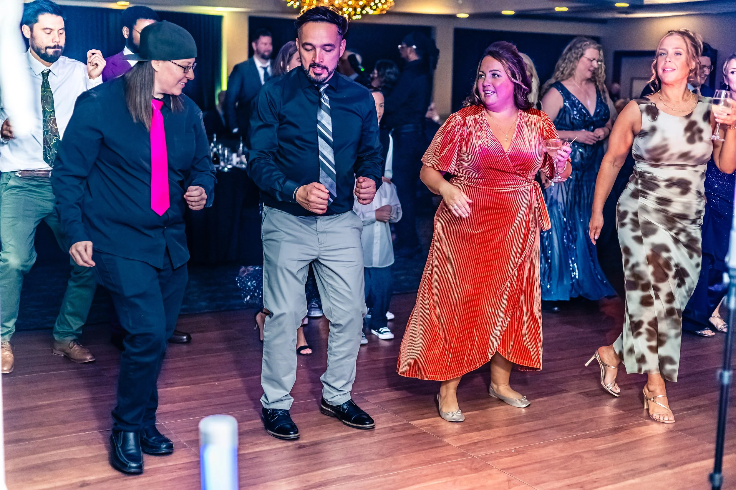 People dancing and enjoying themselves at a party or celebration in an indoor venue, with some holding drinks and wearing formal or semi-formal attire.