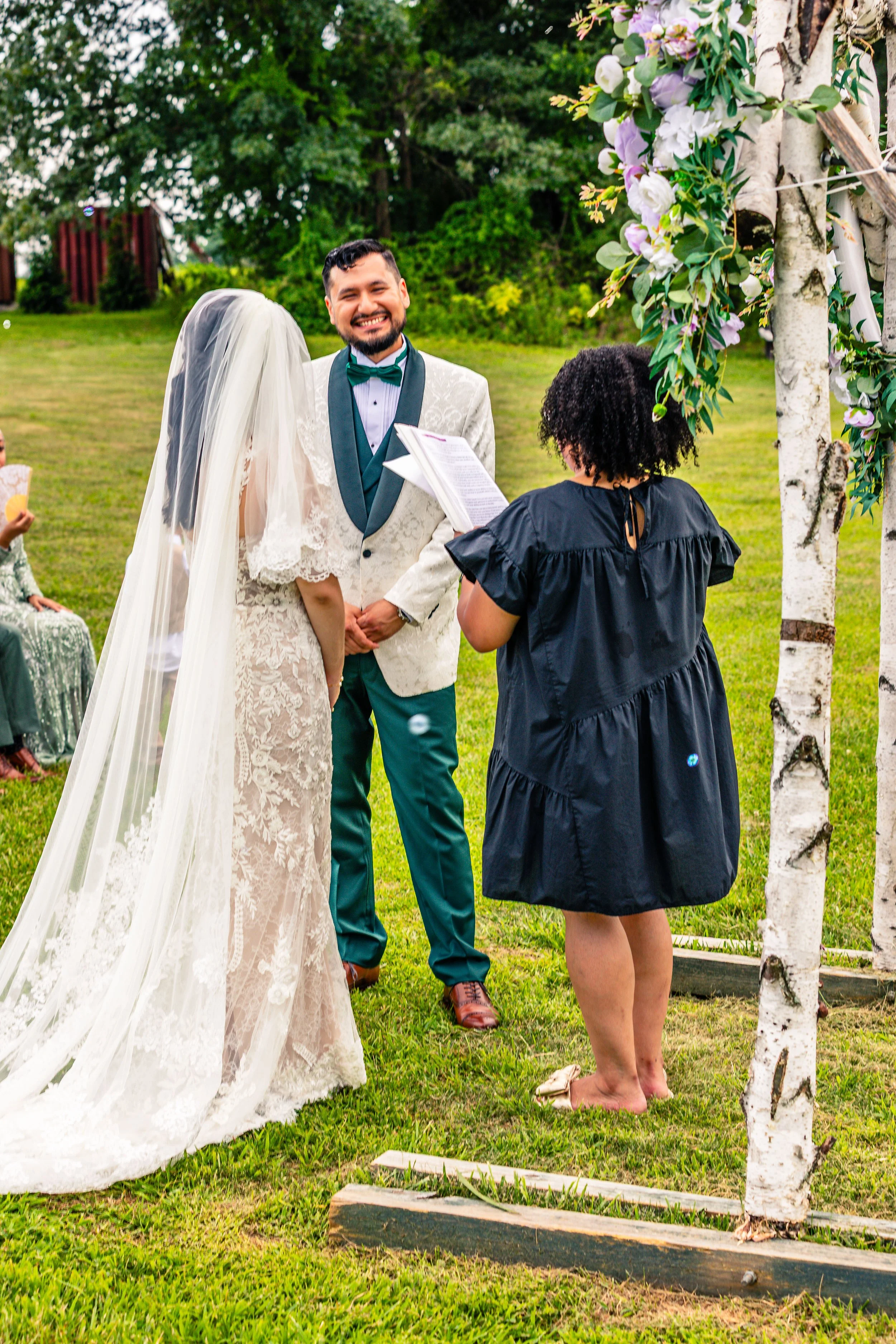 A bride and groom stand outdoors during their wedding ceremony, facing each other and the officiant, with guests seated nearby on a lush green lawn and trees in the background.