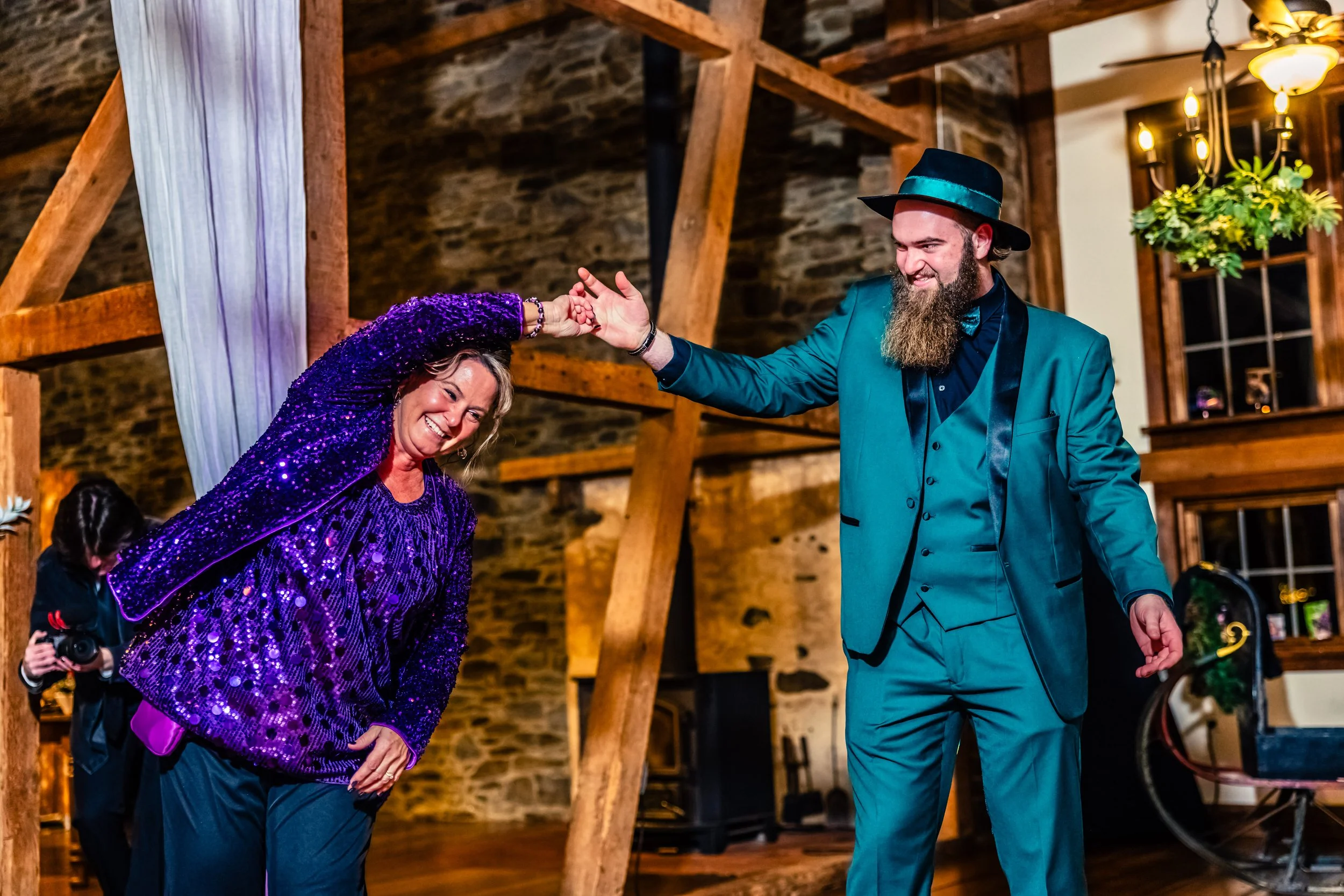 A joyful woman dancing with a bearded man in a teal suit and hat at a rustic, warmly lit indoor event.