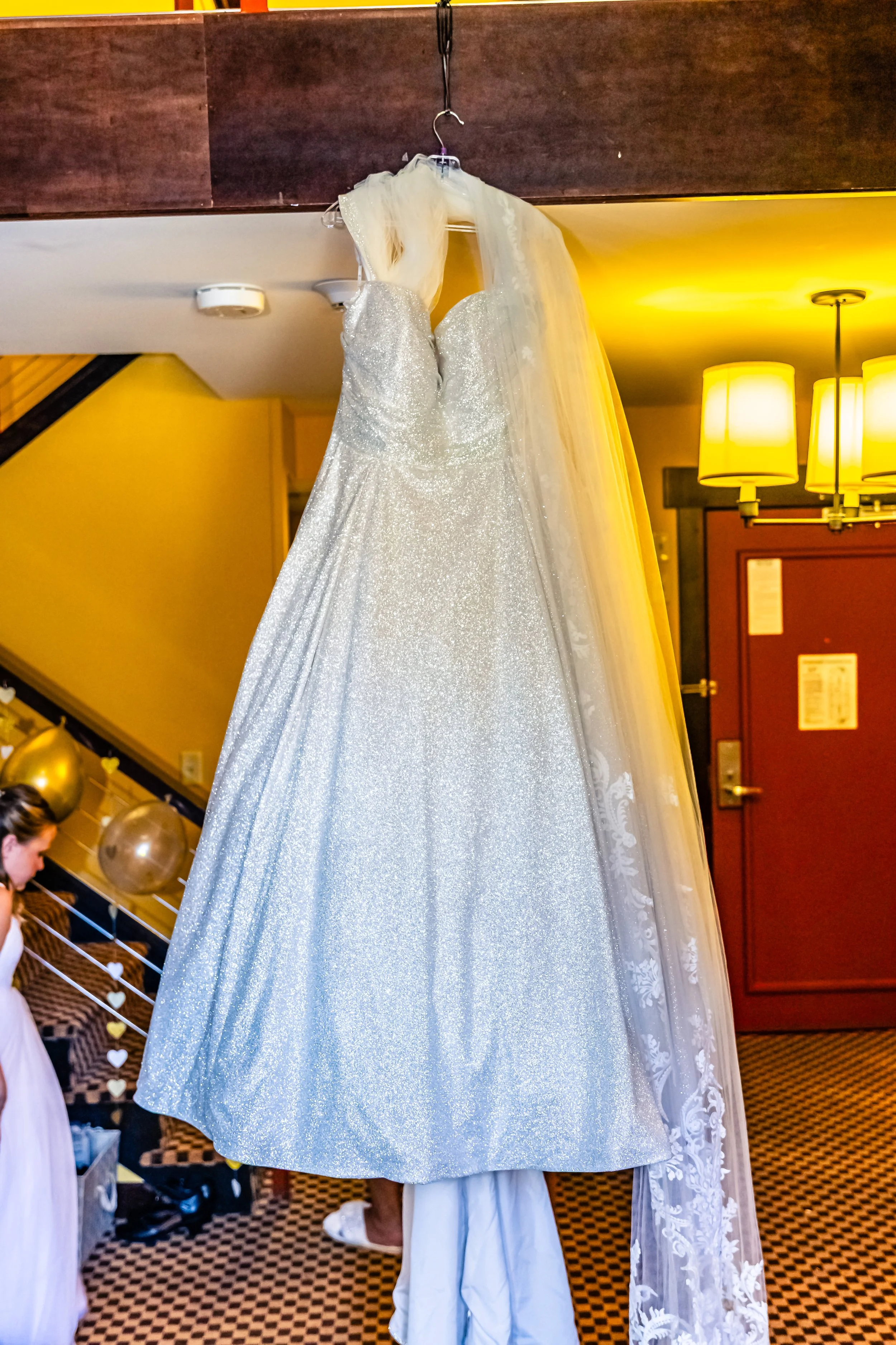 A sparkly white wedding dress hangs on a hanger from a wooden beam, with a lace veil draped over it in a warmly lit room.