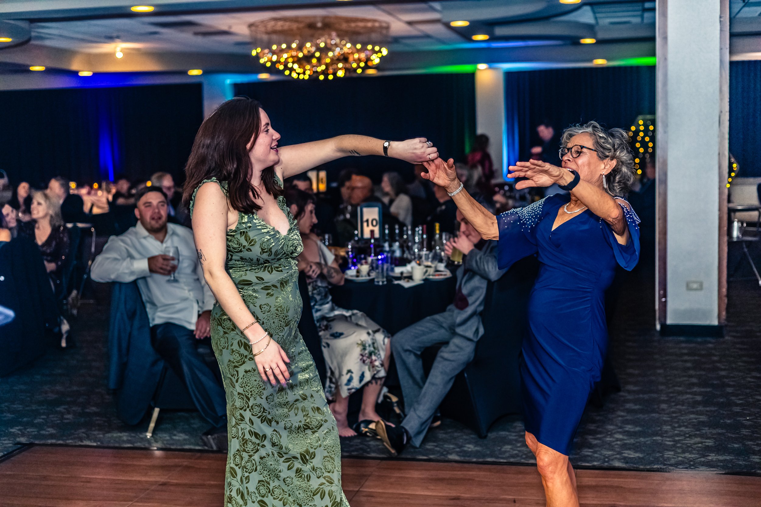 A woman in a blue dress and an older woman in a green dress dancing and playful singing at a party or wedding reception with guests seated at tables in the background.