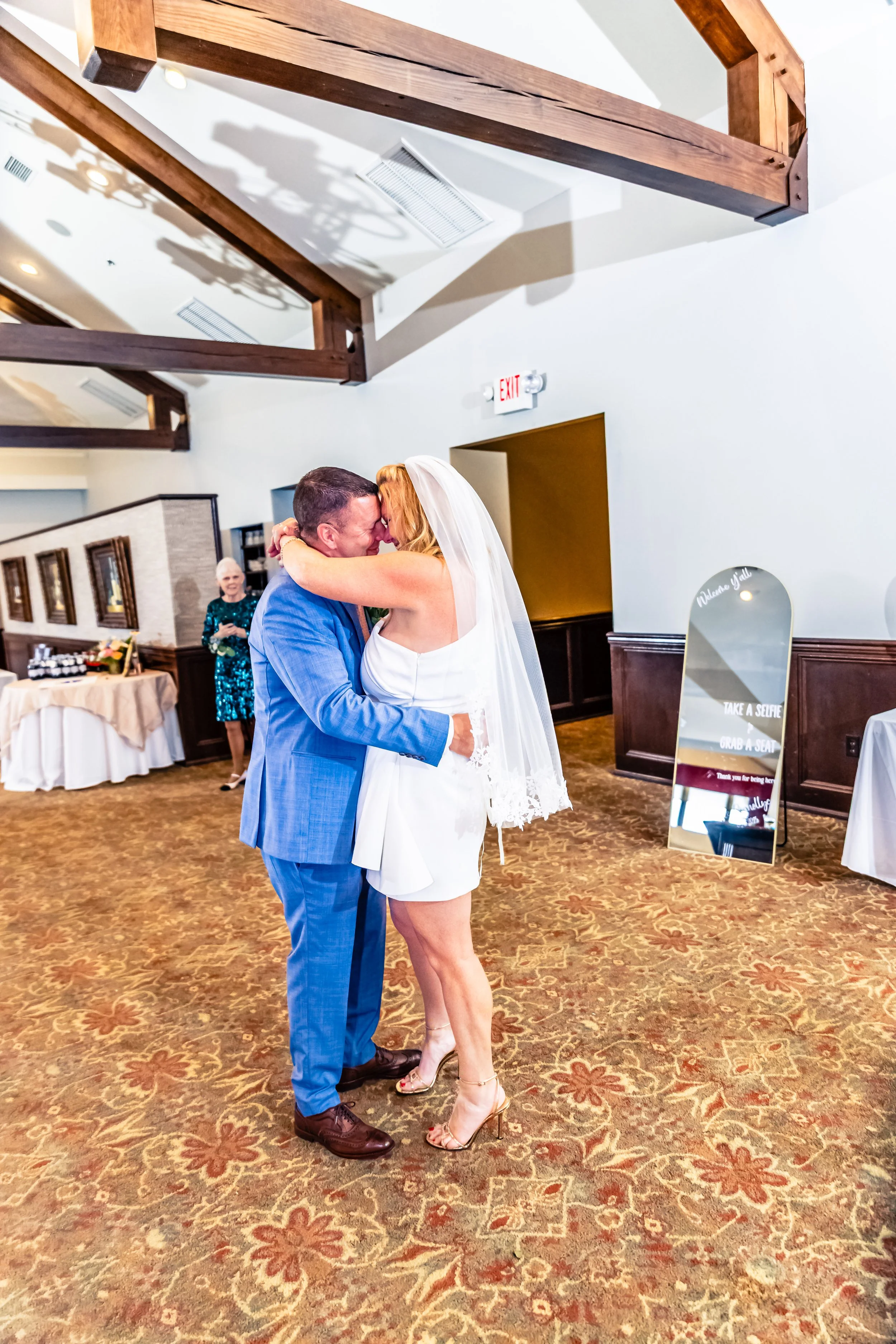 A bride and groom embrace during their wedding reception in a decorated hall, with a woman standing nearby in the background.