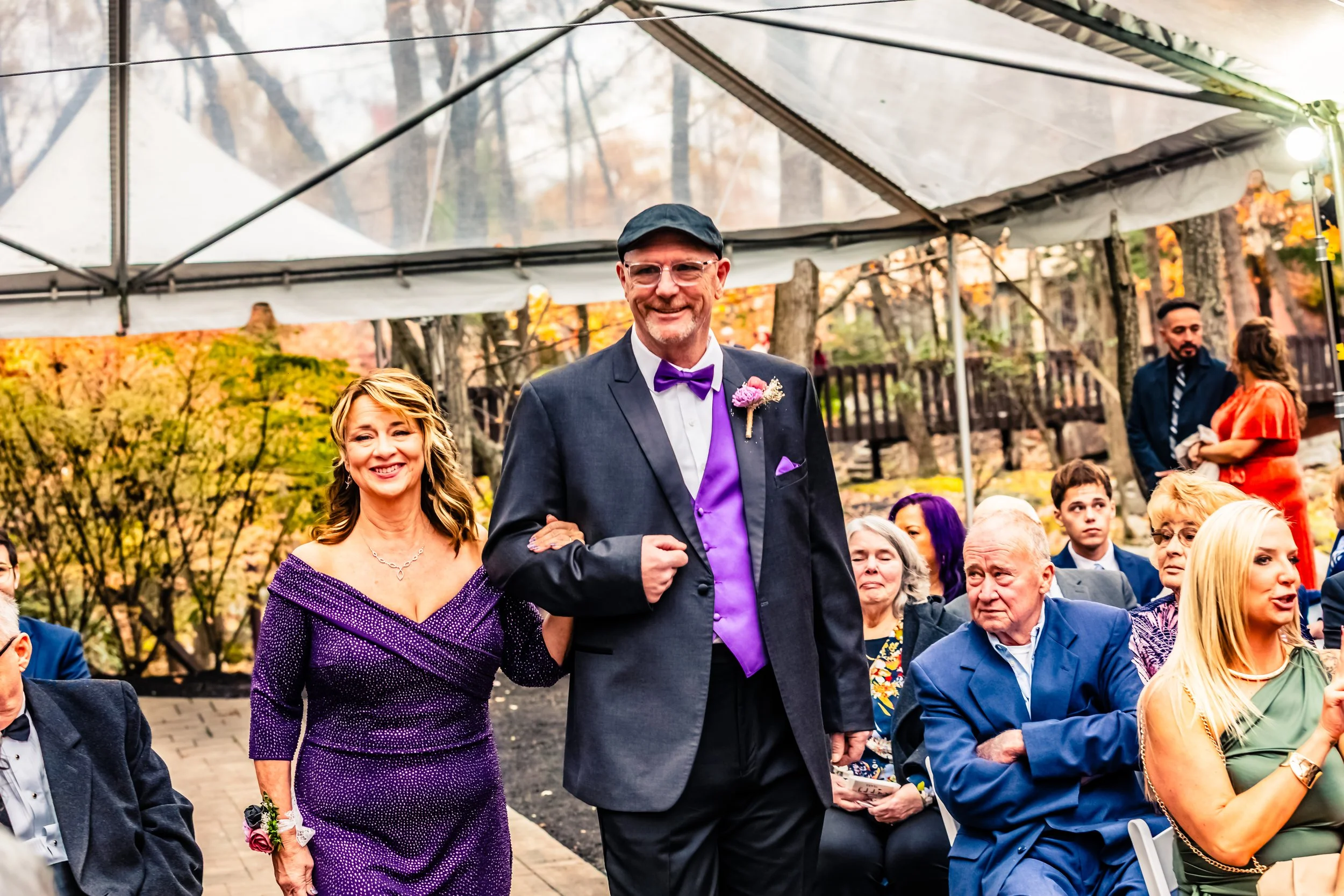 A smiling woman in a purple dress walking arm-in-arm with a smiling man in a suit and purple accents, at an outdoor wedding ceremony with guests seated around them.