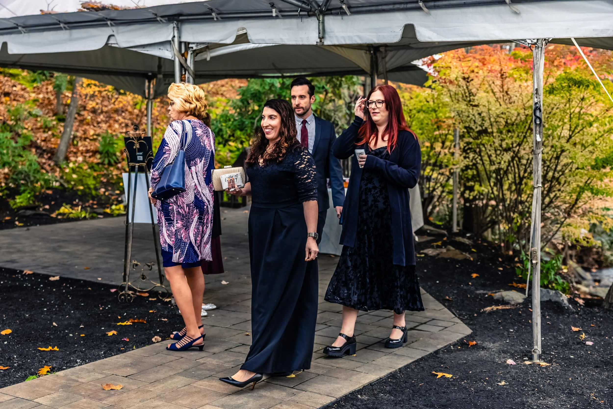 Group of women and a man dressed in formal attire standing outside under a tent, with autumn foliage in the background.