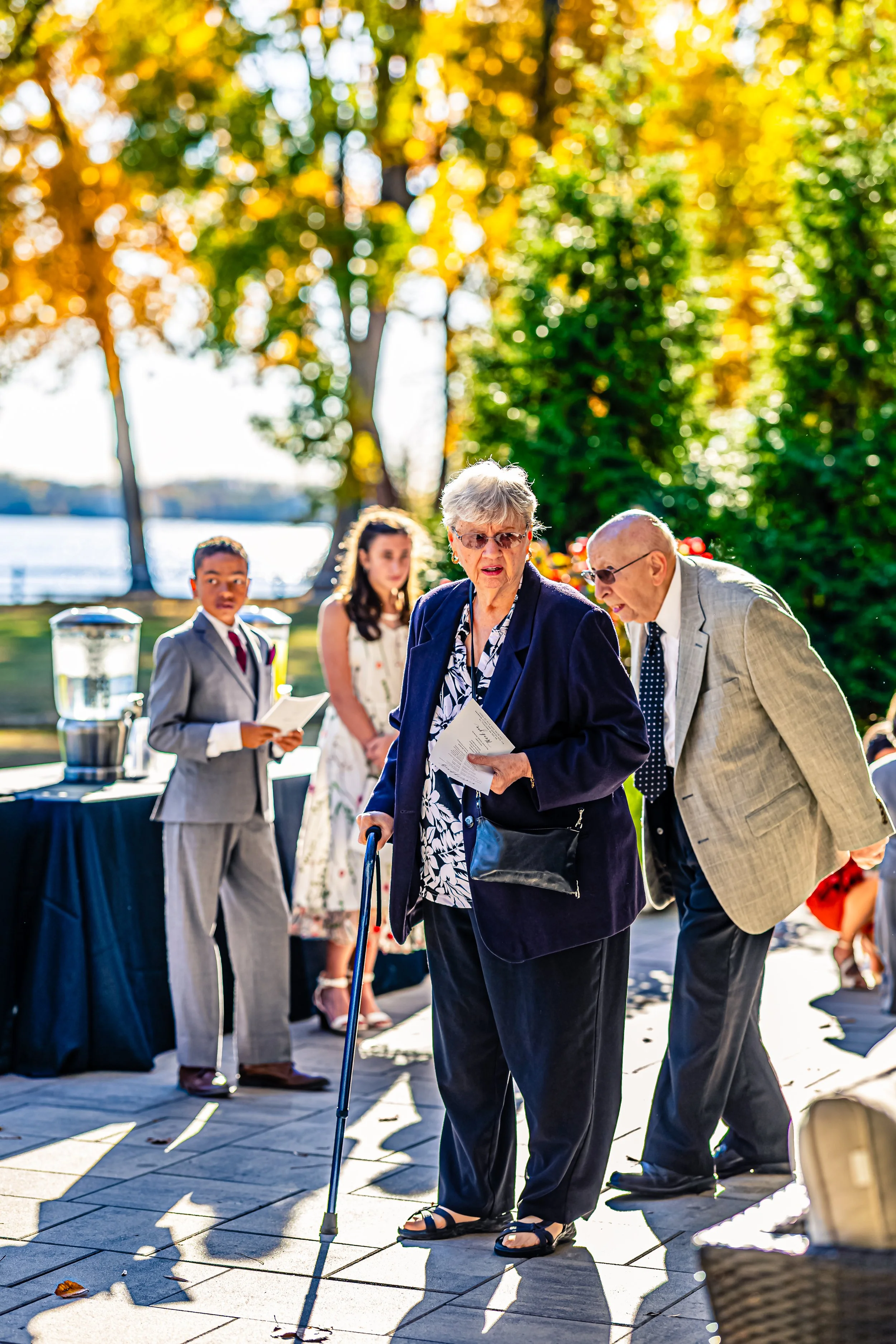 Senior woman with a cane holding papers, and man leaning in with glasses, while two younger people stand nearby at an outdoor event with trees and water in the background.