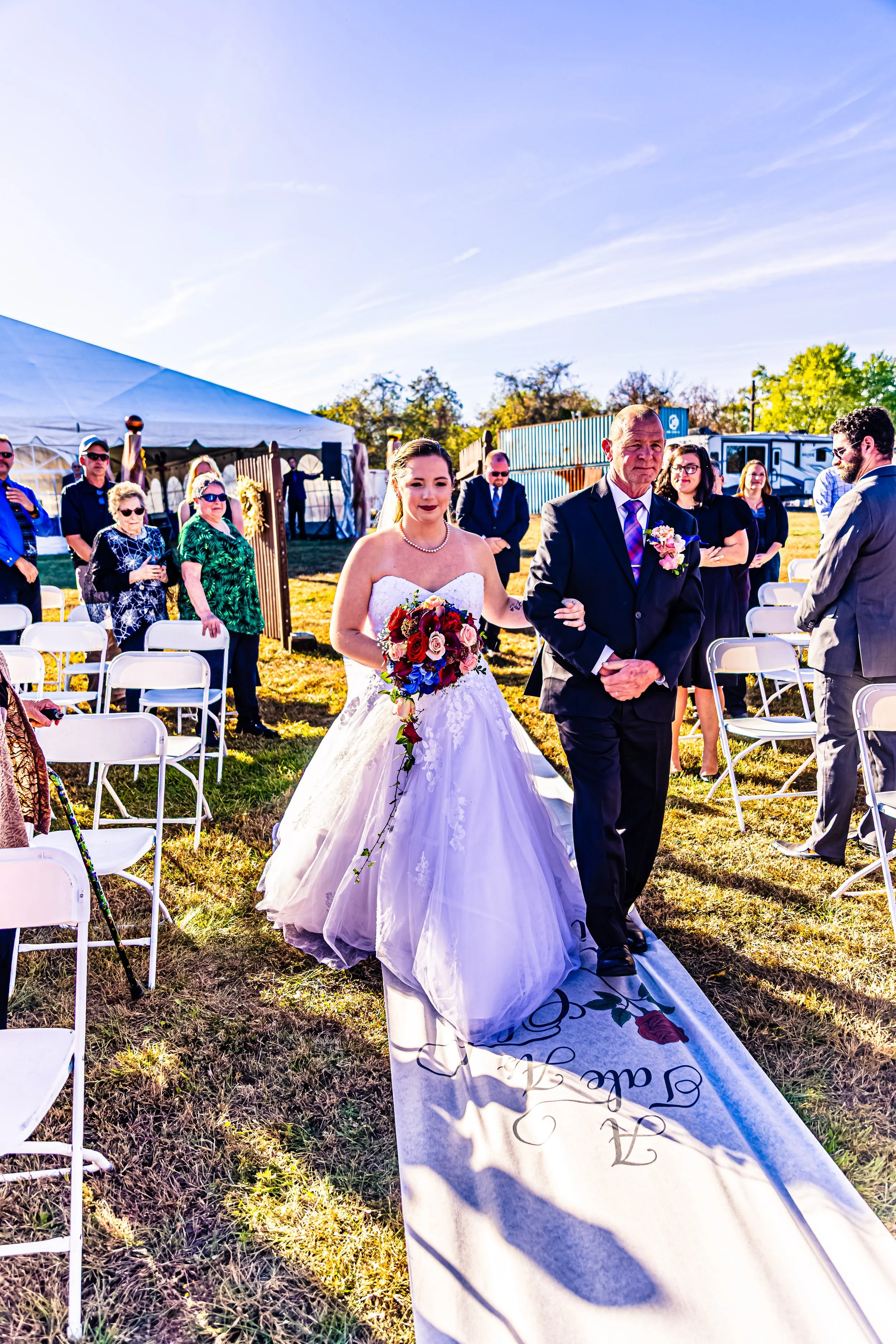 A bride walking down an outdoor aisle, being escorted by a man, at a wedding ceremony with guests seated on either side on a sunny day.