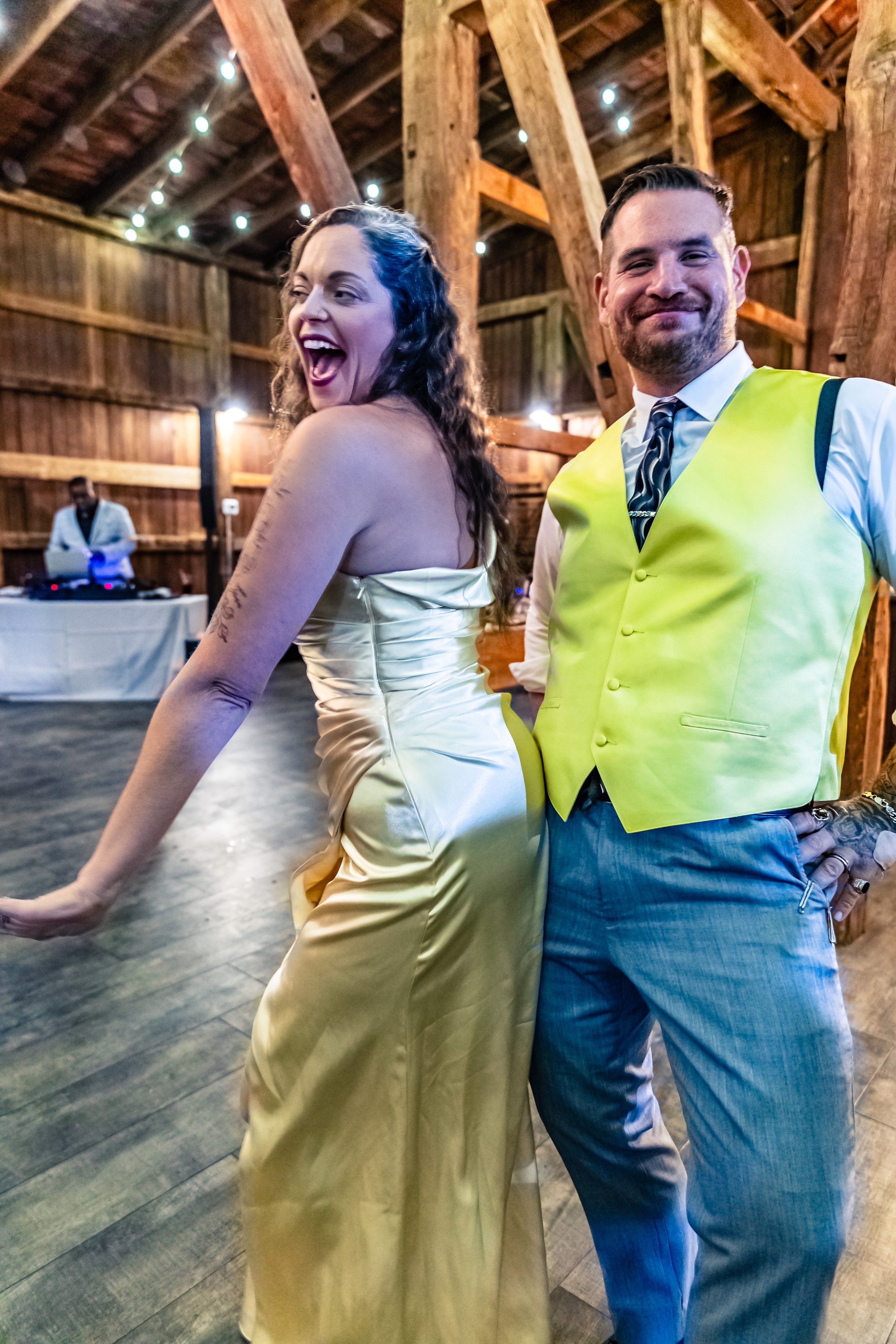 A woman in a shiny gold dress dancing and smiling alongside a man in a white shirt, striped tie, and yellow vest, at a rustic wooden venue with string lights overhead.