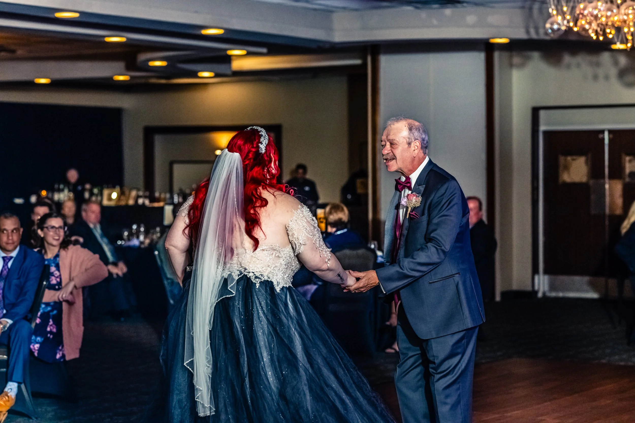 A bride with red hair and a veil dances with an older man in a dark suit at a wedding reception, surrounded by seated guests.
