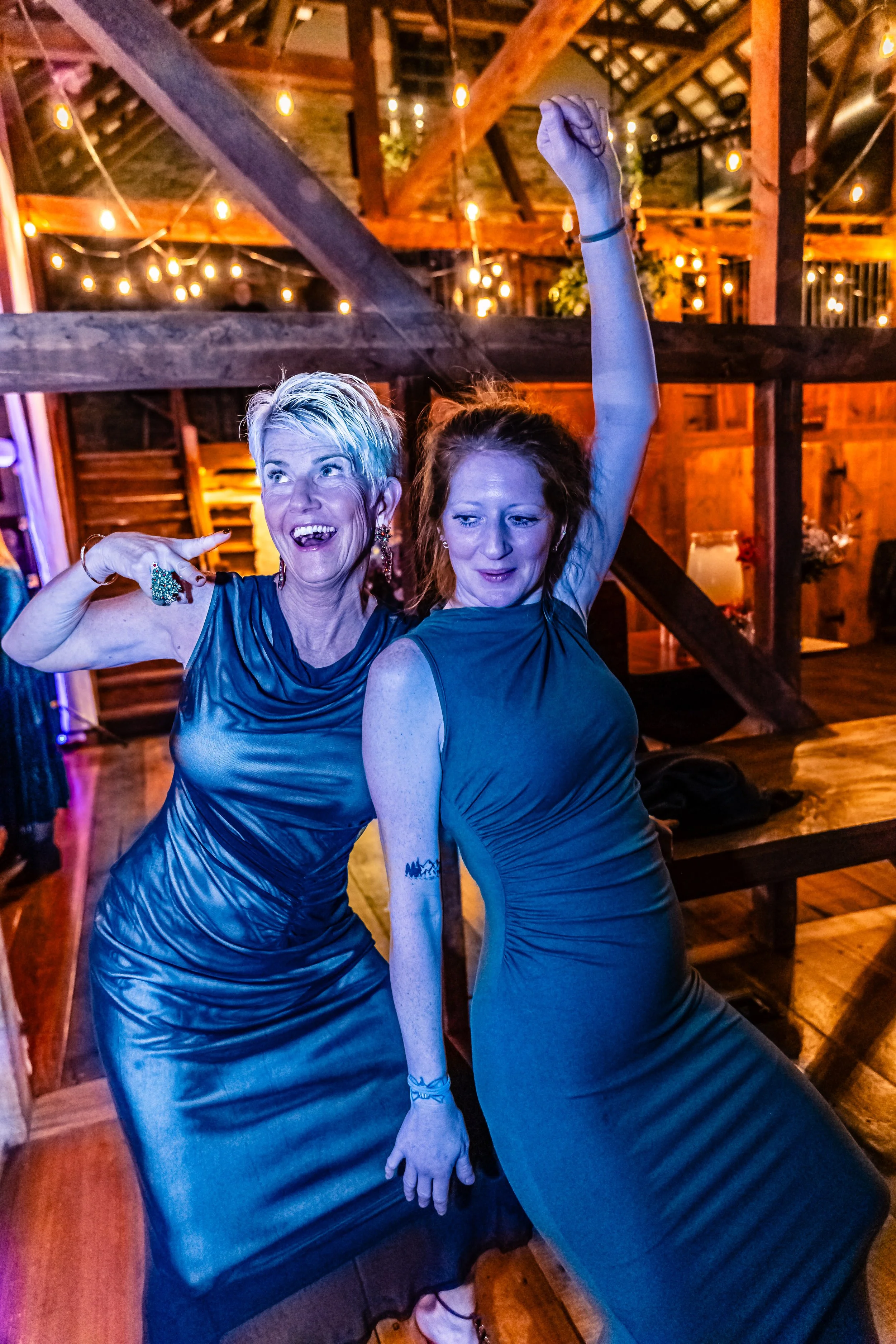 Two women in evening dresses dancing and celebrating at a party with warm, string lights in a rustic venue.