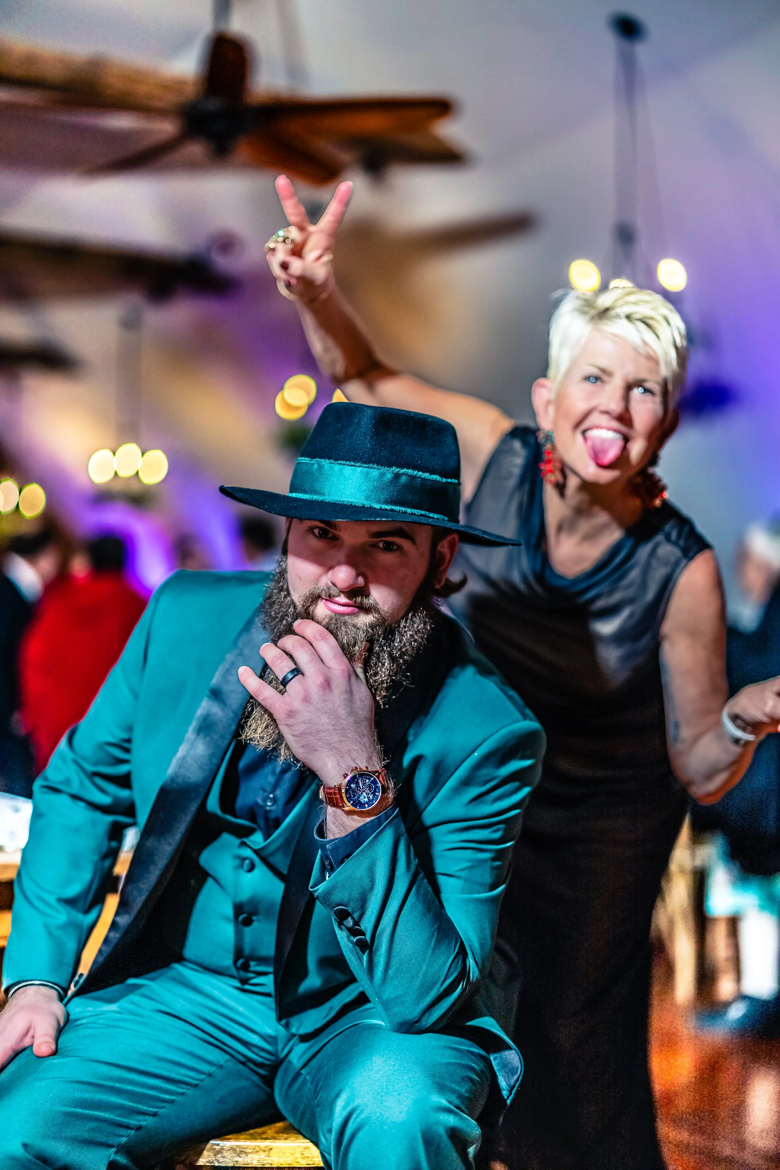 A man with a beard in a teal suit and black hat poses with his hand on his chin, and a woman with short blonde hair, making a playful face and sticking out her tongue, stands behind him at a lively party.