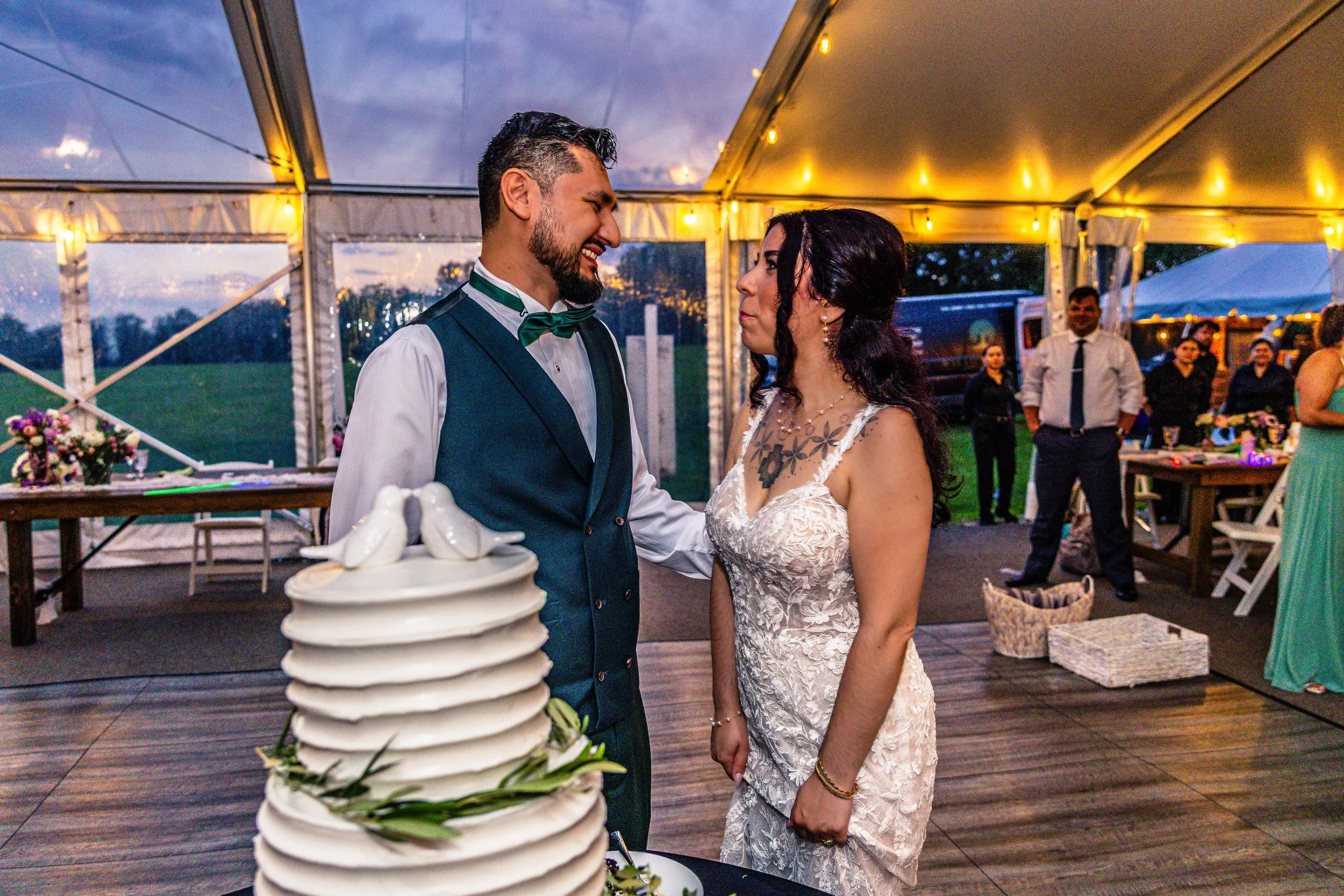 A bride and groom sharing a moment at their wedding reception, with a wedding cake in the foreground and guests in the background under a decorated outdoor tent at sunset.