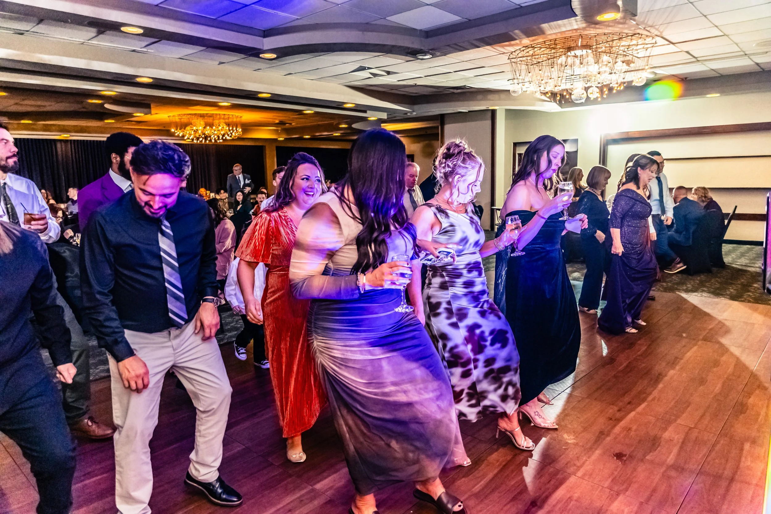 People dancing at a party or celebration in a banquet hall, some holding drinks, with a chandelier and tables in the background.
