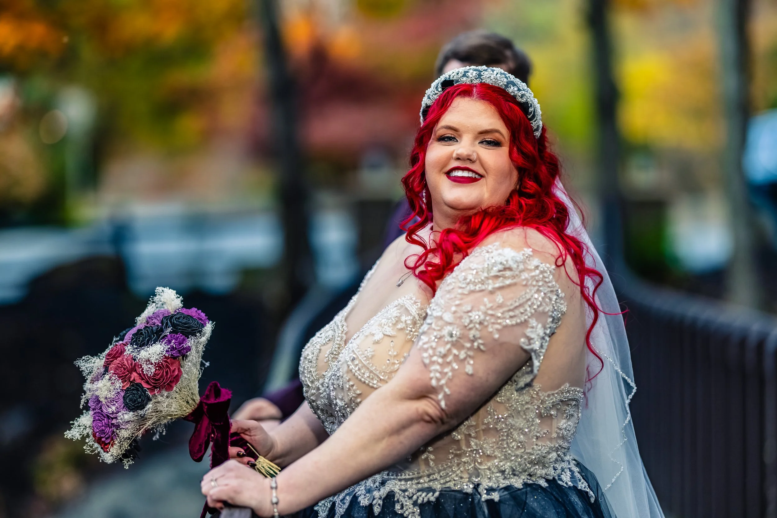 A bride with vibrant red, curly hair, wearing a lace wedding gown with intricate beading, smiling and holding a bouquet of pink, purple, and black flowers, outdoors during fall.