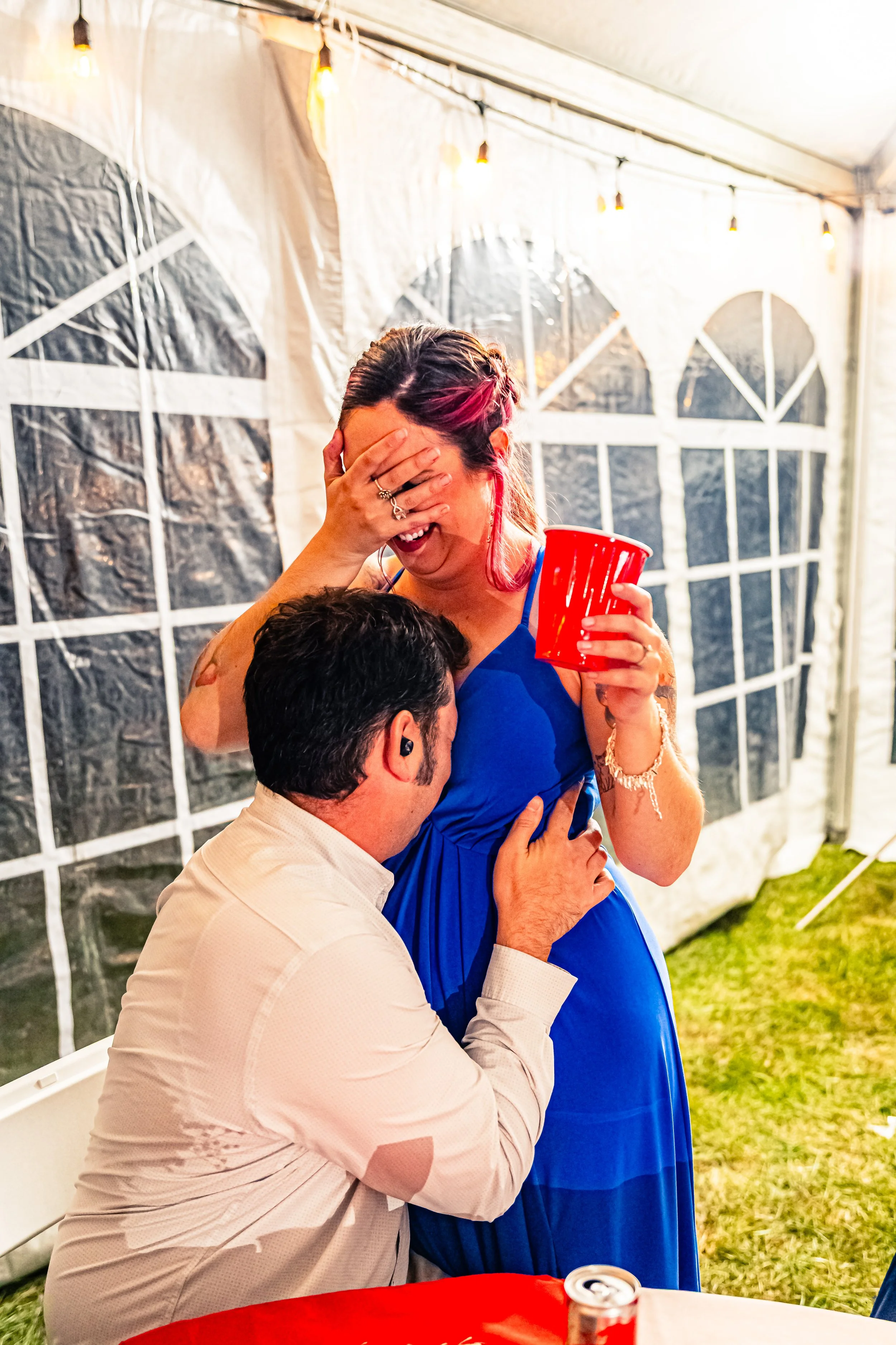 A woman in a blue dress laughing and covering her face with her hand while a man kneels in front of her inside a tent at a celebration, with a red cup in her hand.
