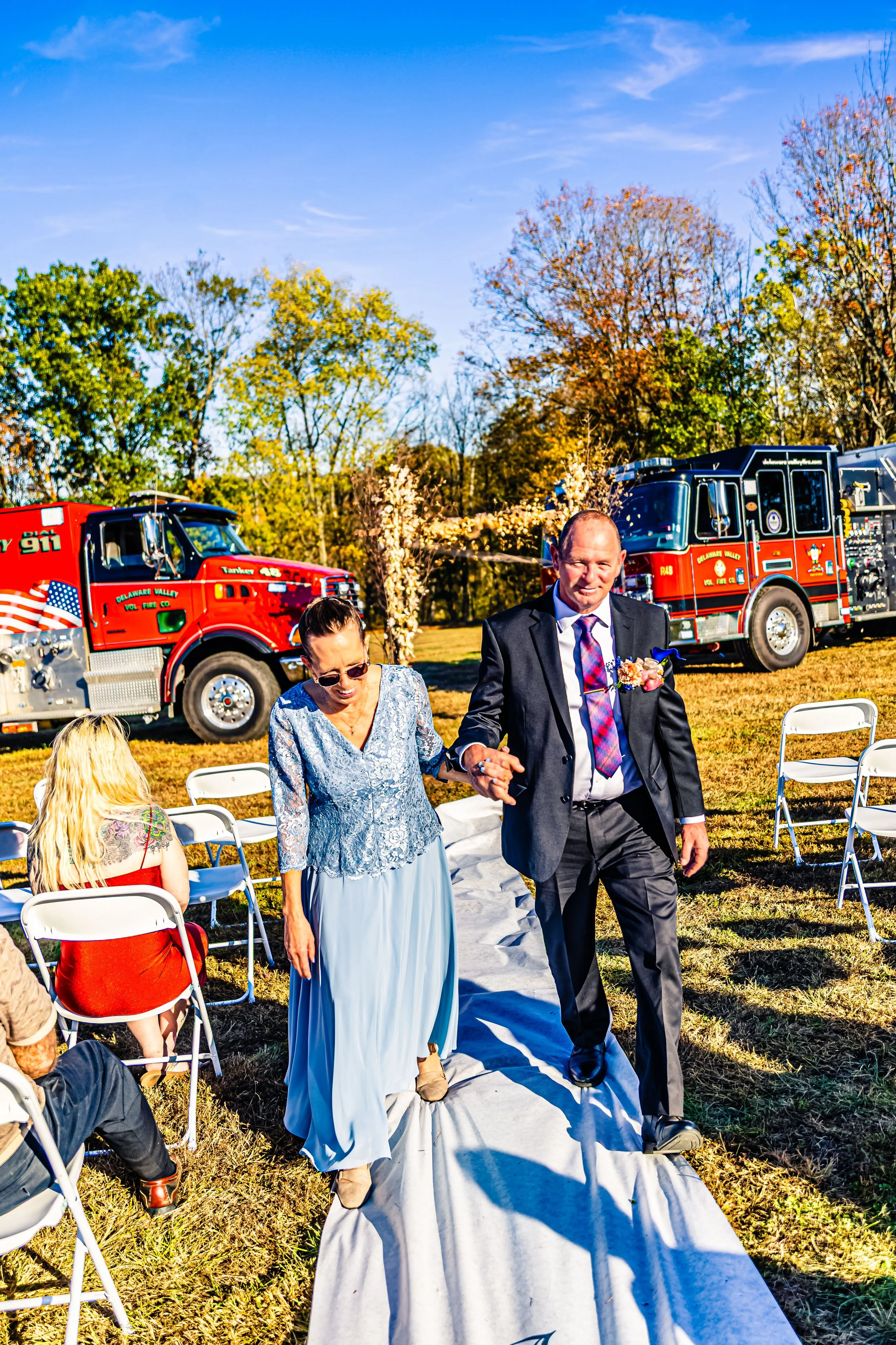A couple walking down a white aisle runner at an outdoor wedding ceremony, with fire trucks in the background and guests seated on white chairs on a grassy field, trees with fall foliage, and a clear blue sky.