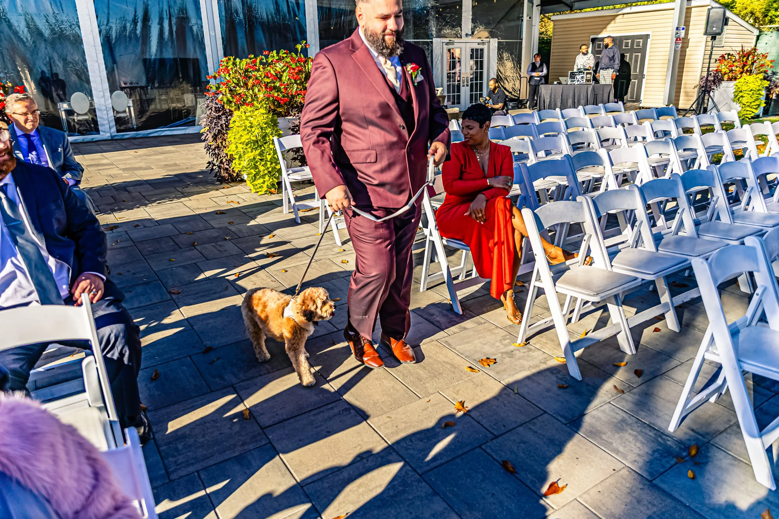 A man in a maroon suit holding a small dog on a leash at an outdoor wedding ceremony. A woman in a red dress is sitting nearby, and other guests are seated, with some standing near a DJ booth in the background. The setting includes white folding chai