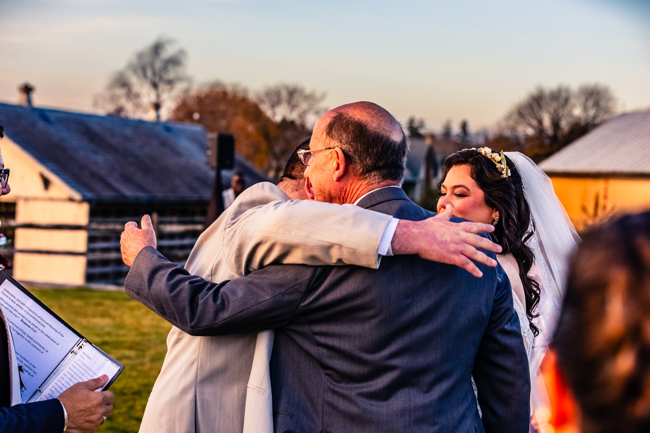 A bride hugging a man, likely her father, during a wedding ceremony outdoors at sunset, with guests and rustic barns in the background.