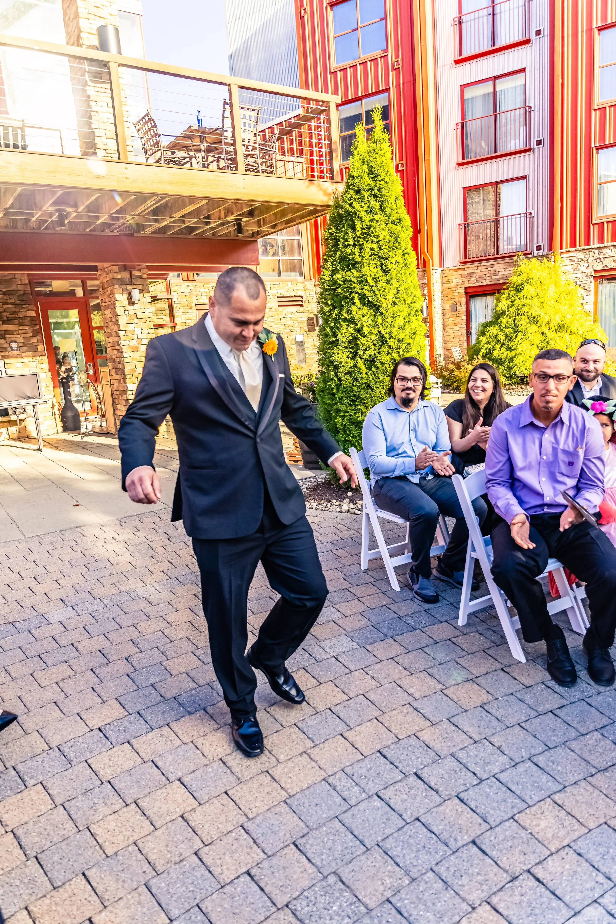 A man in a dark suit and white shirt dancing outdoors at a wedding or formal event, with seated guests clapping and smiling in the background, in a colorful urban courtyard setting.
