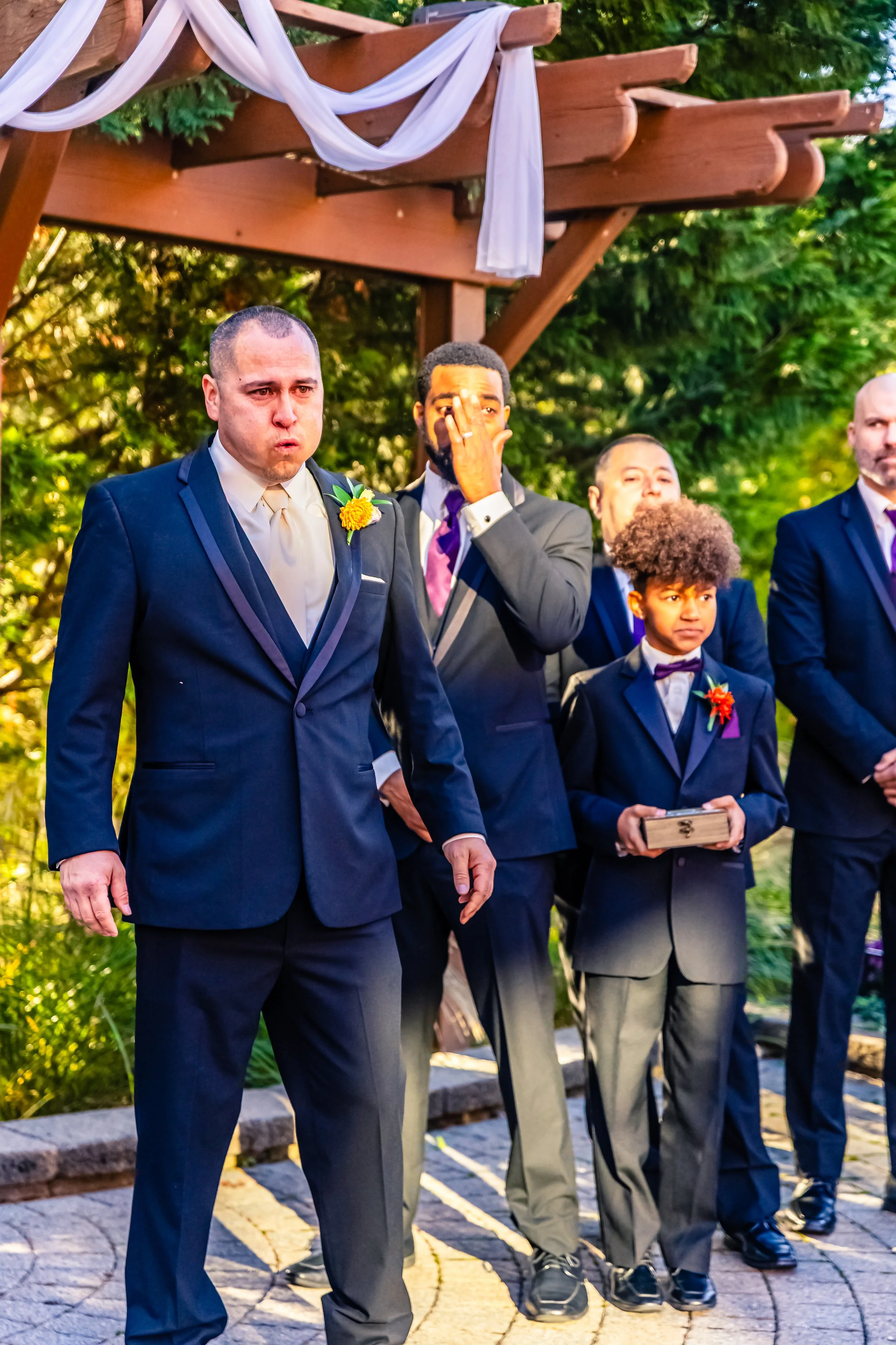 Group of men and a boy dressed in formal suits standing outdoors during a wedding ceremony.