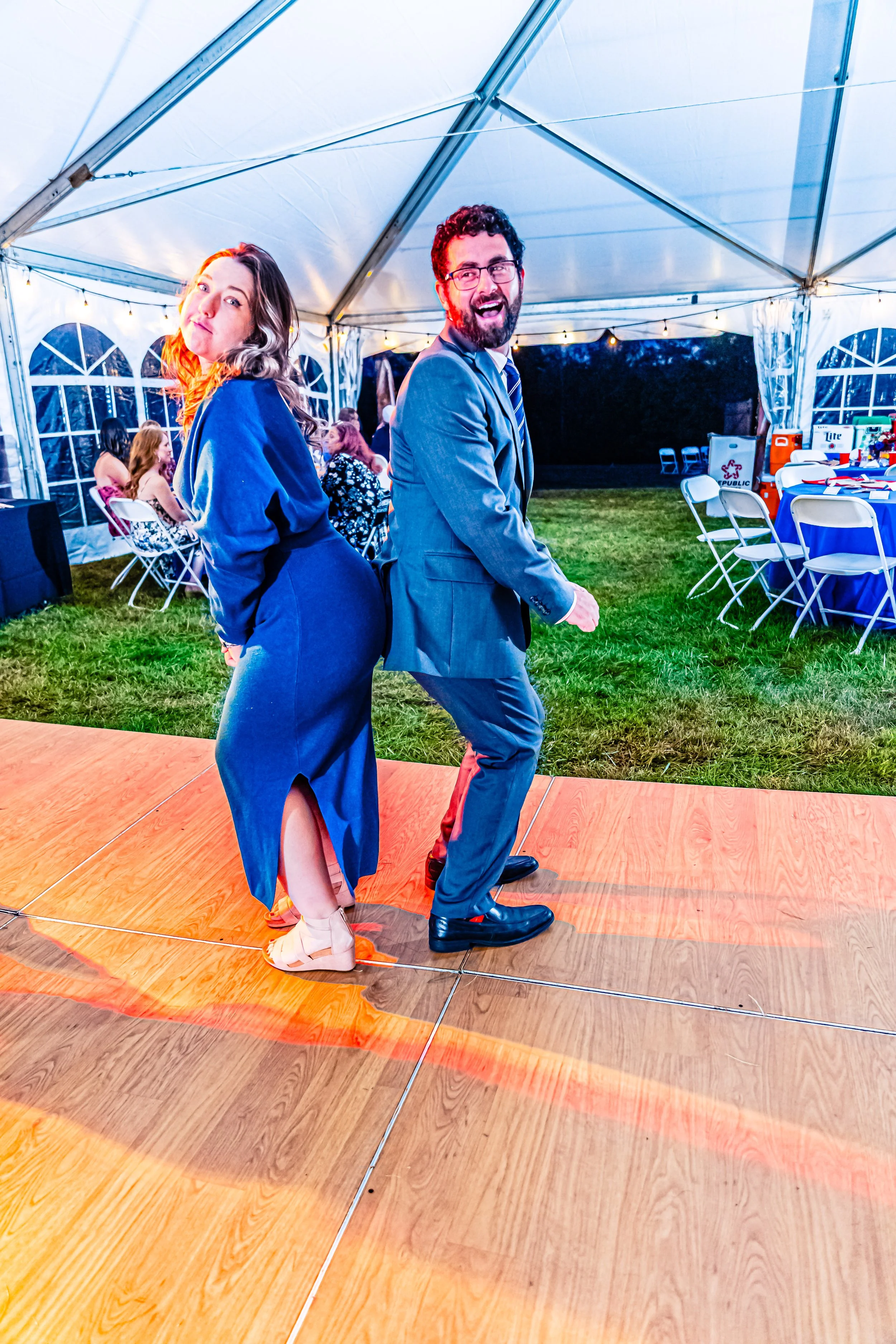 A man and woman dancing back-to-back on a wooden dance floor at an outdoor event tent with tables and guests in the background.