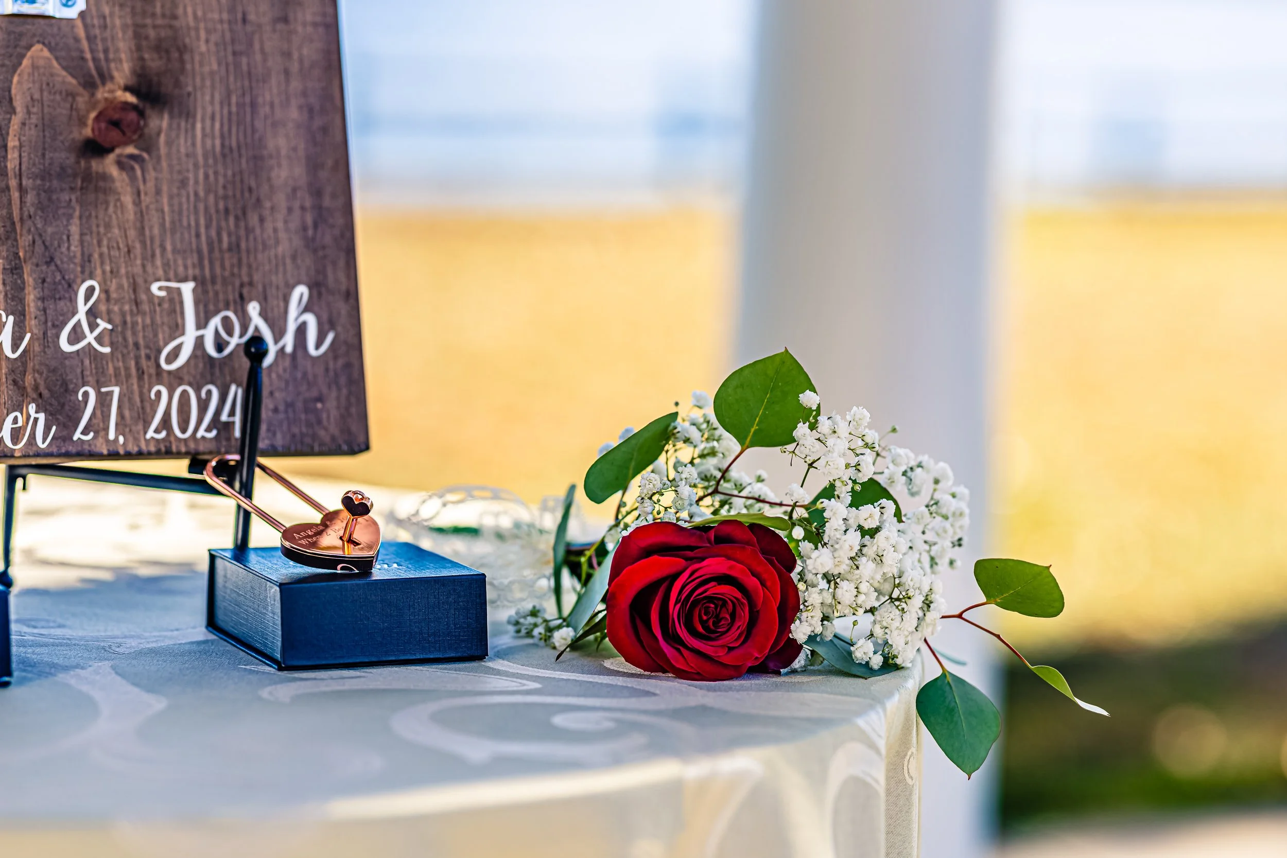 A wedding table with a floral arrangement of a red rose, white baby's breath, and green leaves, a wooden sign with the names  & Josh, and a small black box with a rose gold key as a wedding favor.