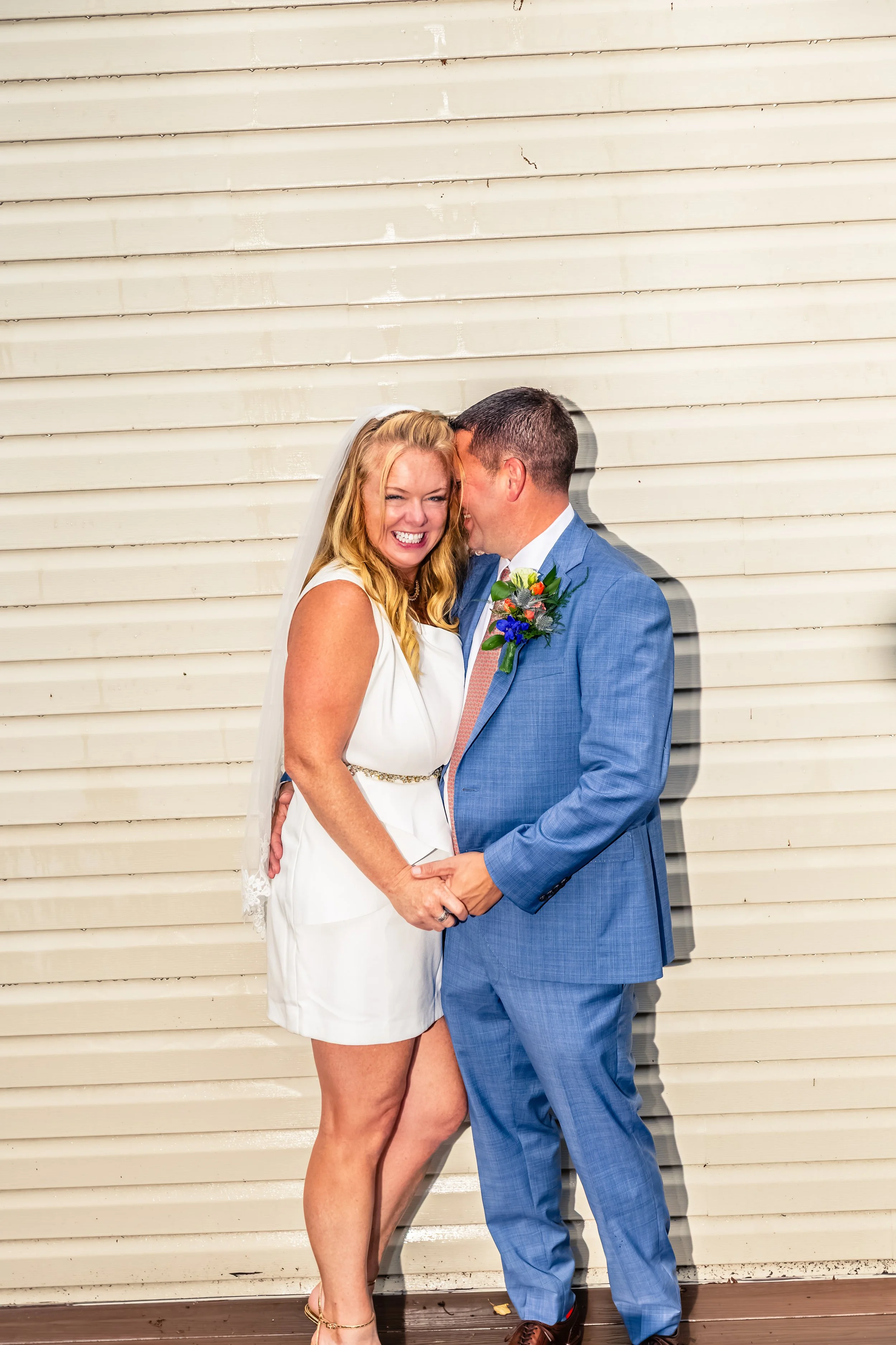 A happy bride in a white dress and a groom in a blue suit smiling and touching foreheads against a beige wall.