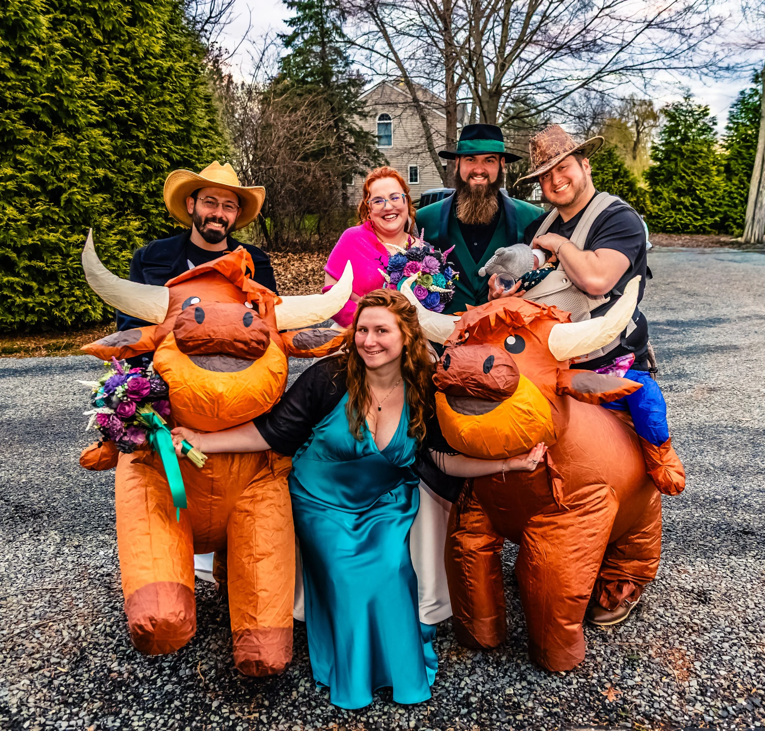 A group of five people celebrating outdoors during a party or wedding, with two inflatable cows decorated with flowers. The group includes three men and two women, with some wearing hats and colorful clothing, posing together in front of a suburban h