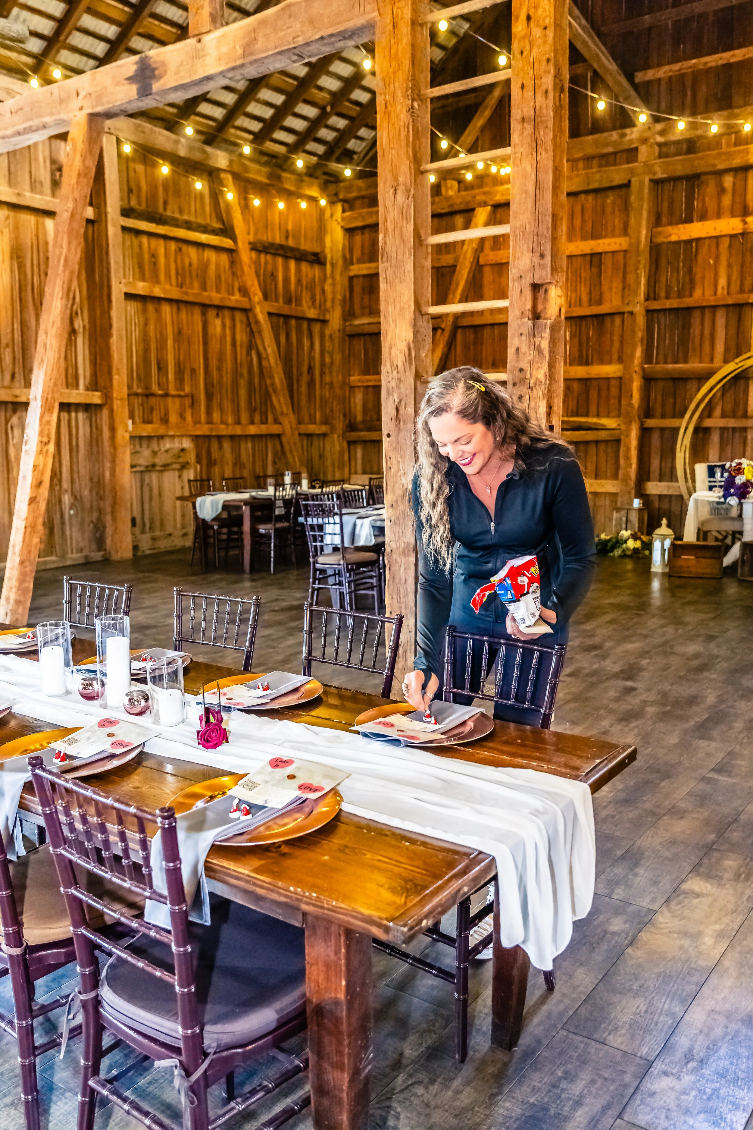 A woman preparing a decorated table in a rustic wooden barn for a celebration, with string lights hanging from the ceiling.