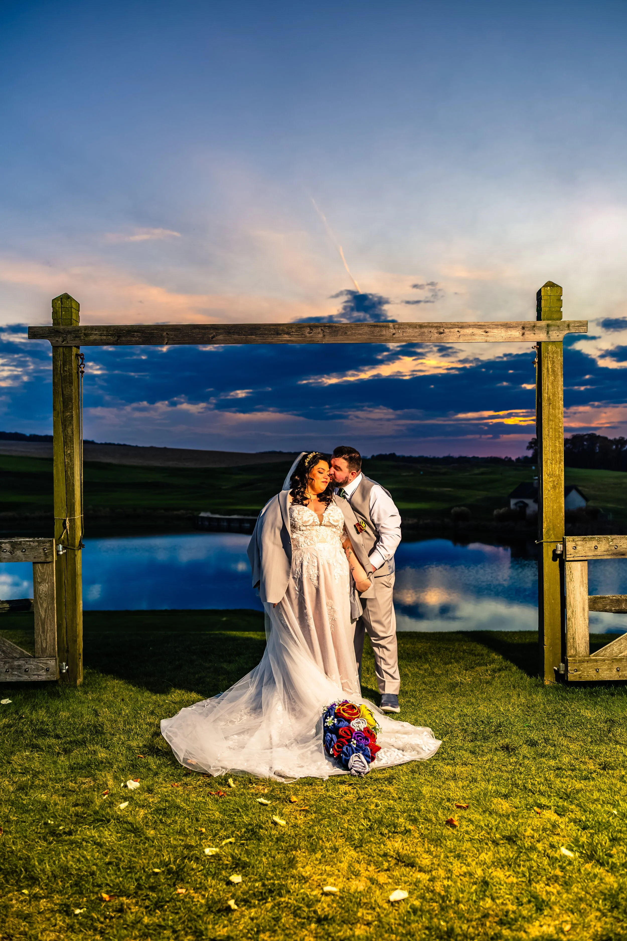 A bride and groom standing together outdoors at sunset, with a lake and rolling hills in the background. The bride is in a white wedding dress, holding a bouquet of colorful flowers, and the groom is in a light-colored suit. They are under a wooden a