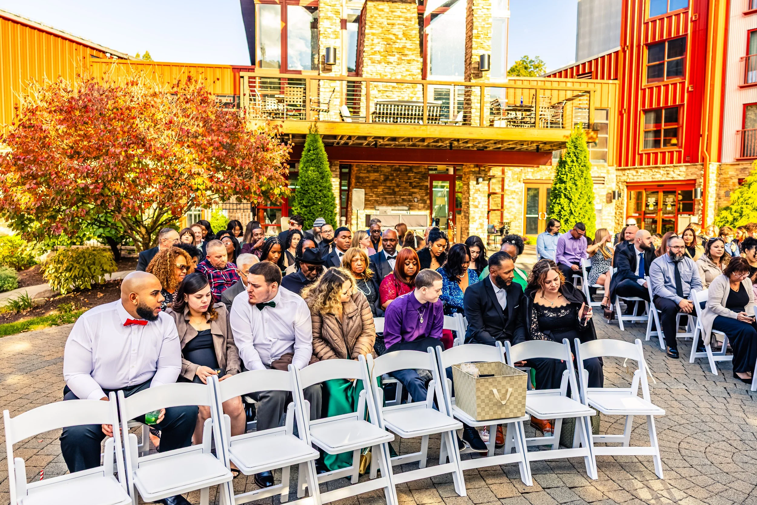 People seated outdoors at a wedding or event, with a colorful building and trees in the background.