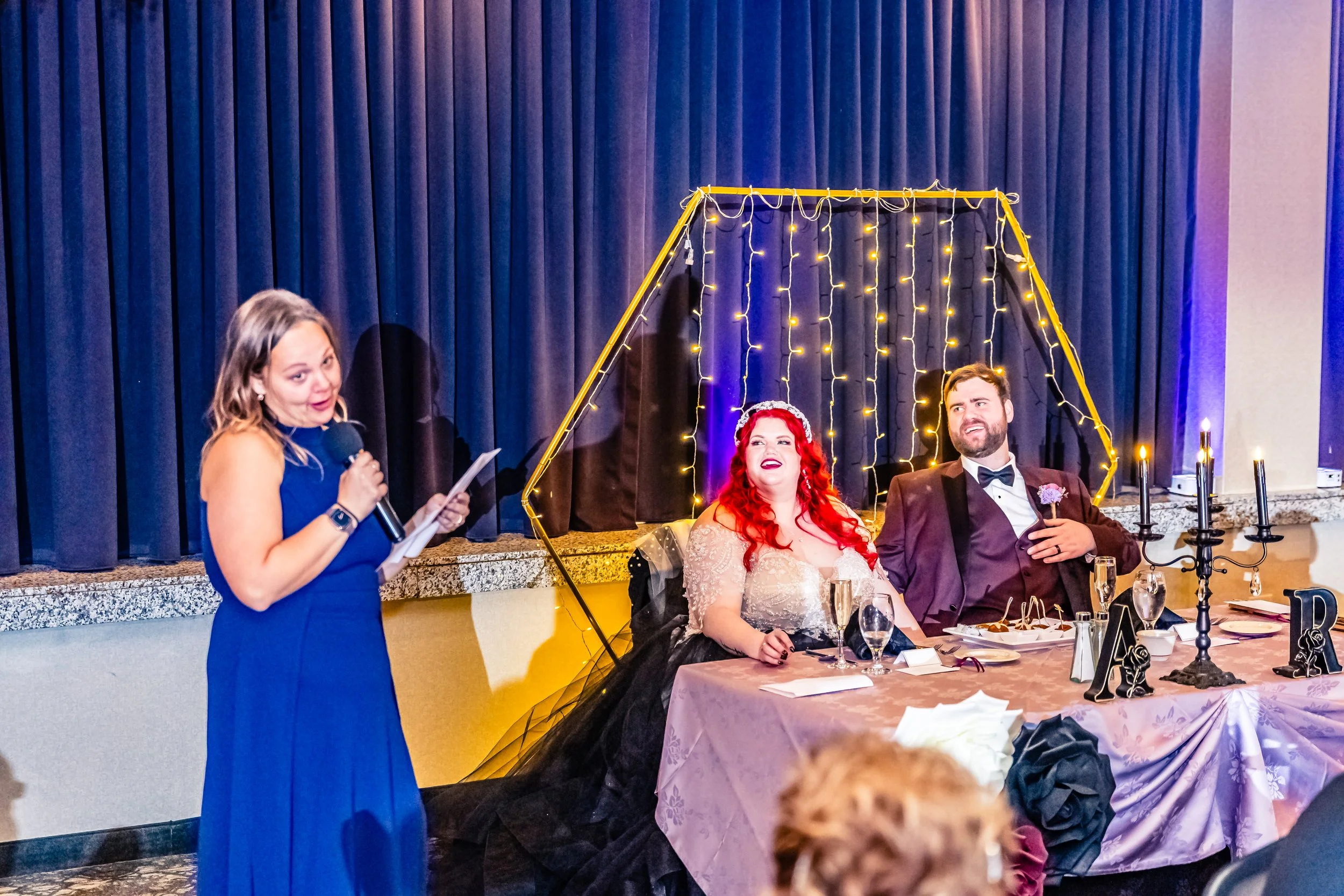 A wedding reception scene with a woman giving a speech, a bride with red hair and a groom sitting at a decorated table behind her, with blue curtains and string lights in the background.