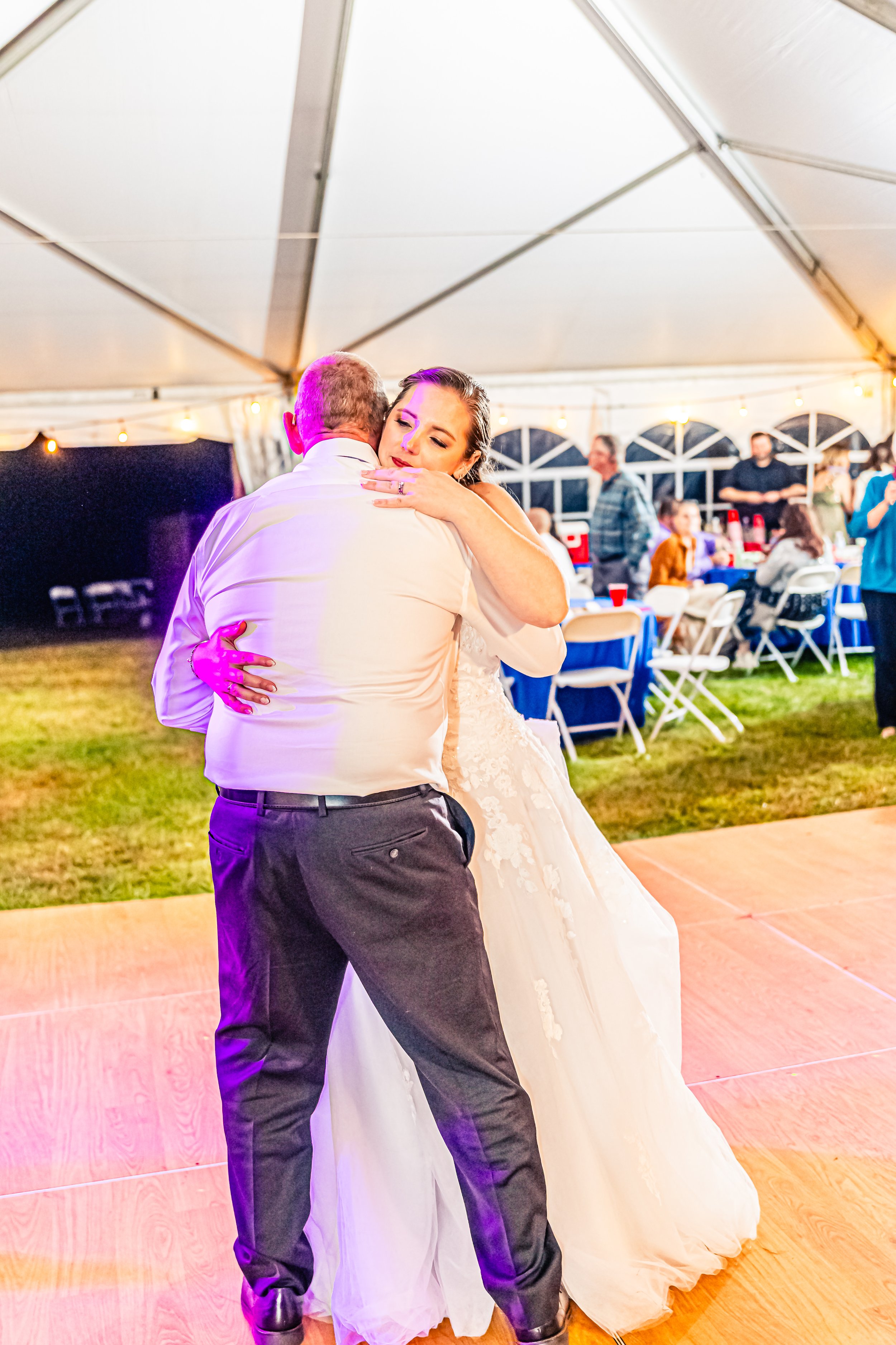 A bride and an older man are dancing closely at a wedding reception under a large white tent, with people seated at tables in the background.