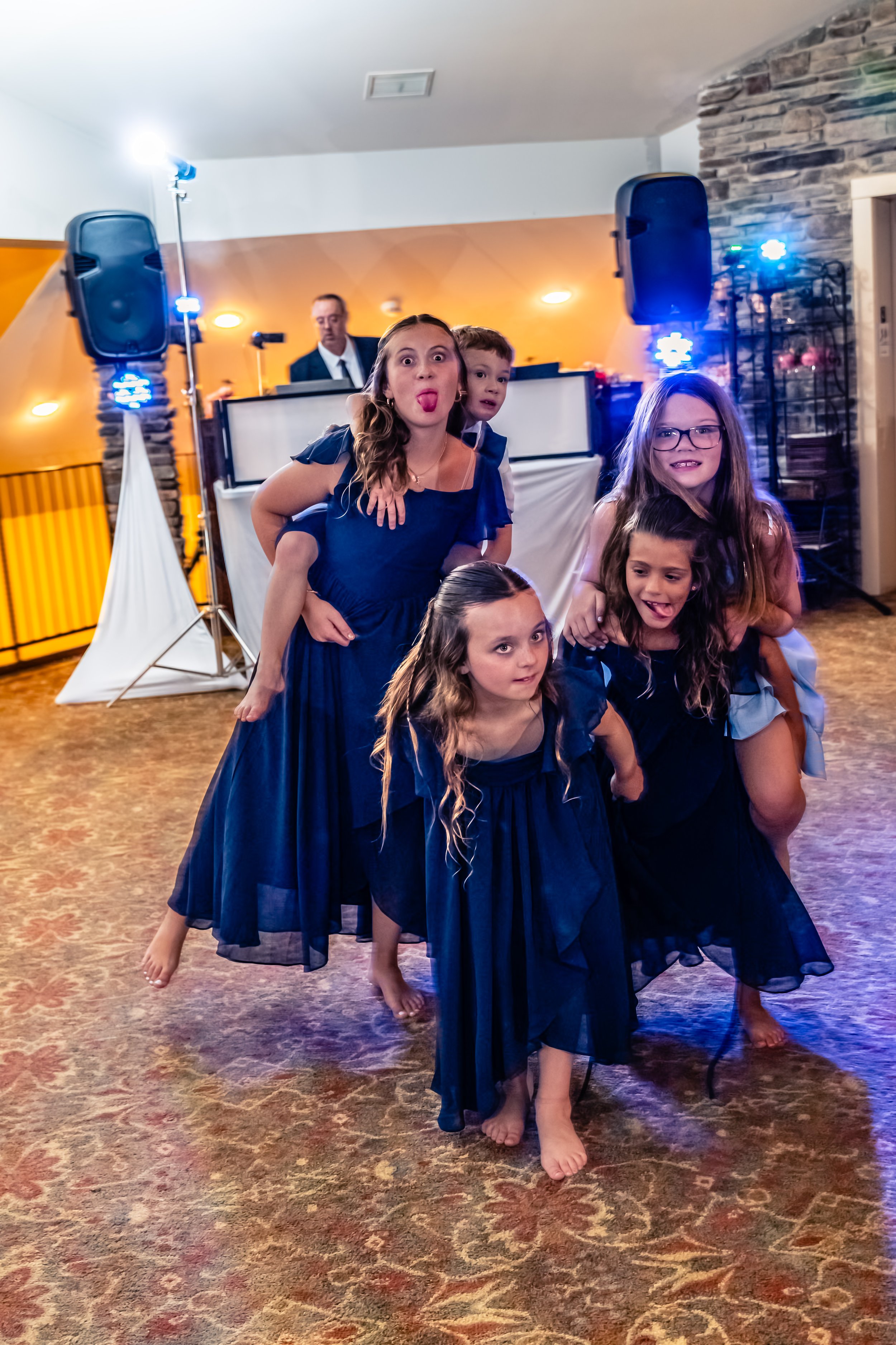 A group of five children with three girls in blue dresses and two boys, gathered in a line at a party, with a DJ and speakers in the background.