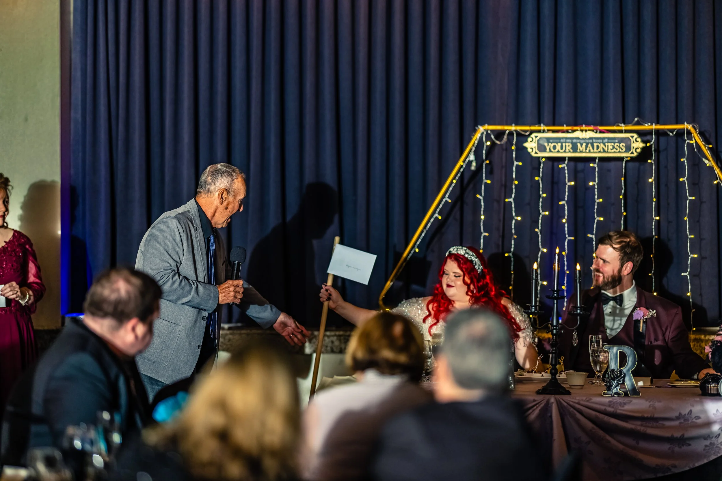 A wedding reception scene with a bride with red hair and a groom sitting at a decorated table, a man in a gray jacket speaking into a microphone, and guests seated at tables. The backdrop features dark curtains and a sign that says "Your Madness."